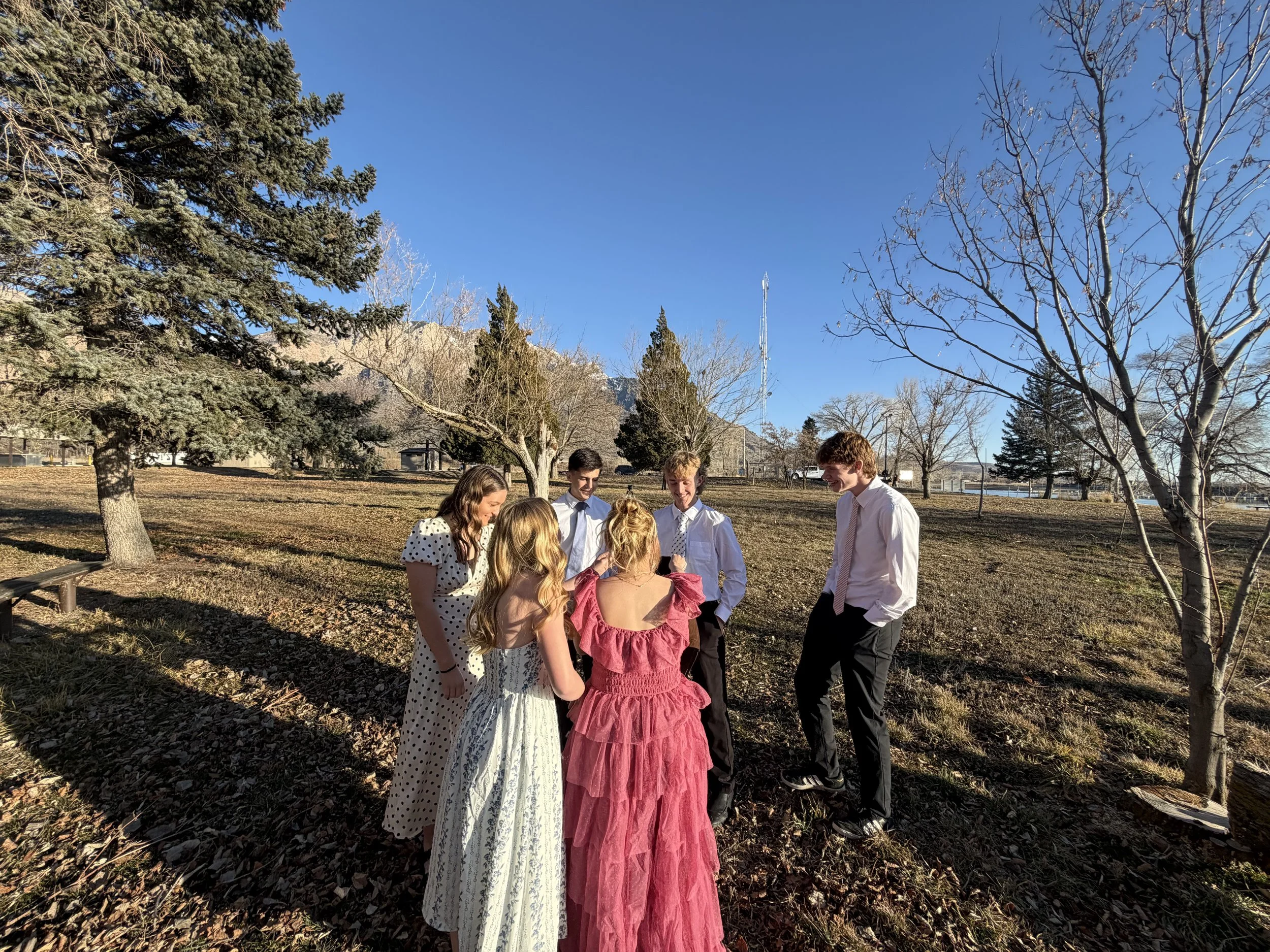 Group of young people dressed in formal attire standing outdoors in a park with trees and a clear blue sky.