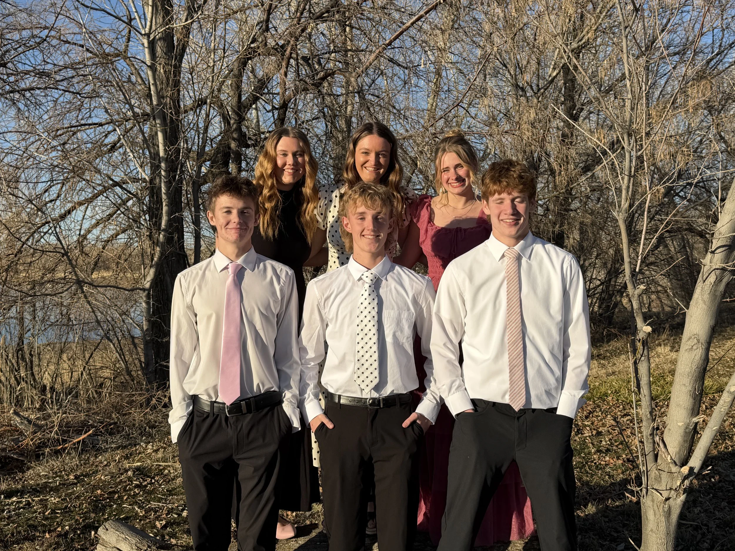 Group of six young people outdoors in front of trees, dressed semi-formally with boys in button-up shirts and ties, girls in dresses, smiling.