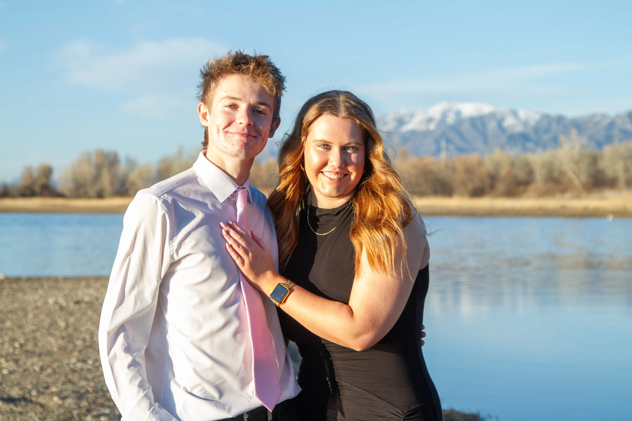A young man and woman standing together by the water on a sunny day, with mountains and trees in the background.