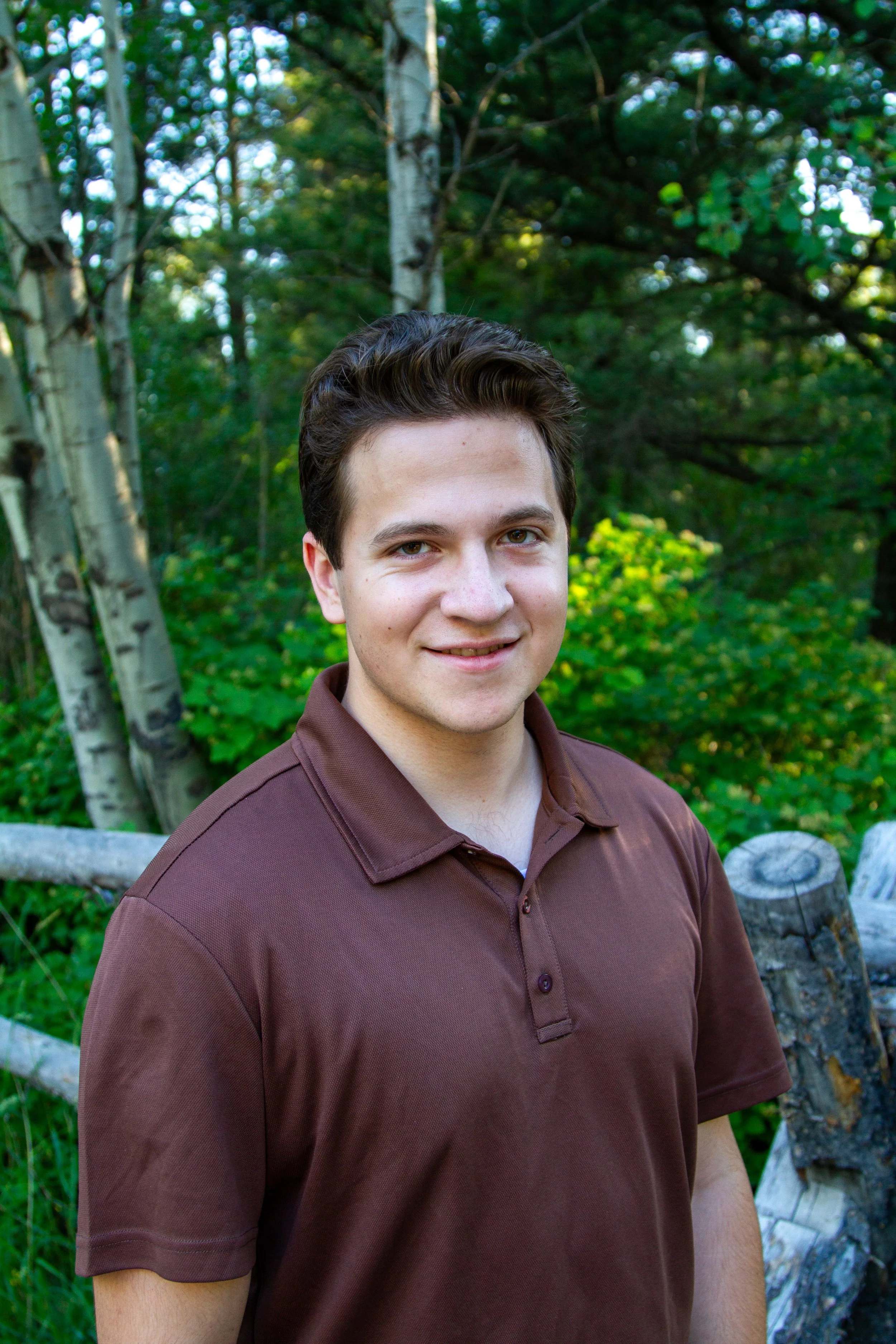 A young man with dark brown hair, wearing a brown polo shirt, standing outdoors in a wooded area with green trees and logs in the background.