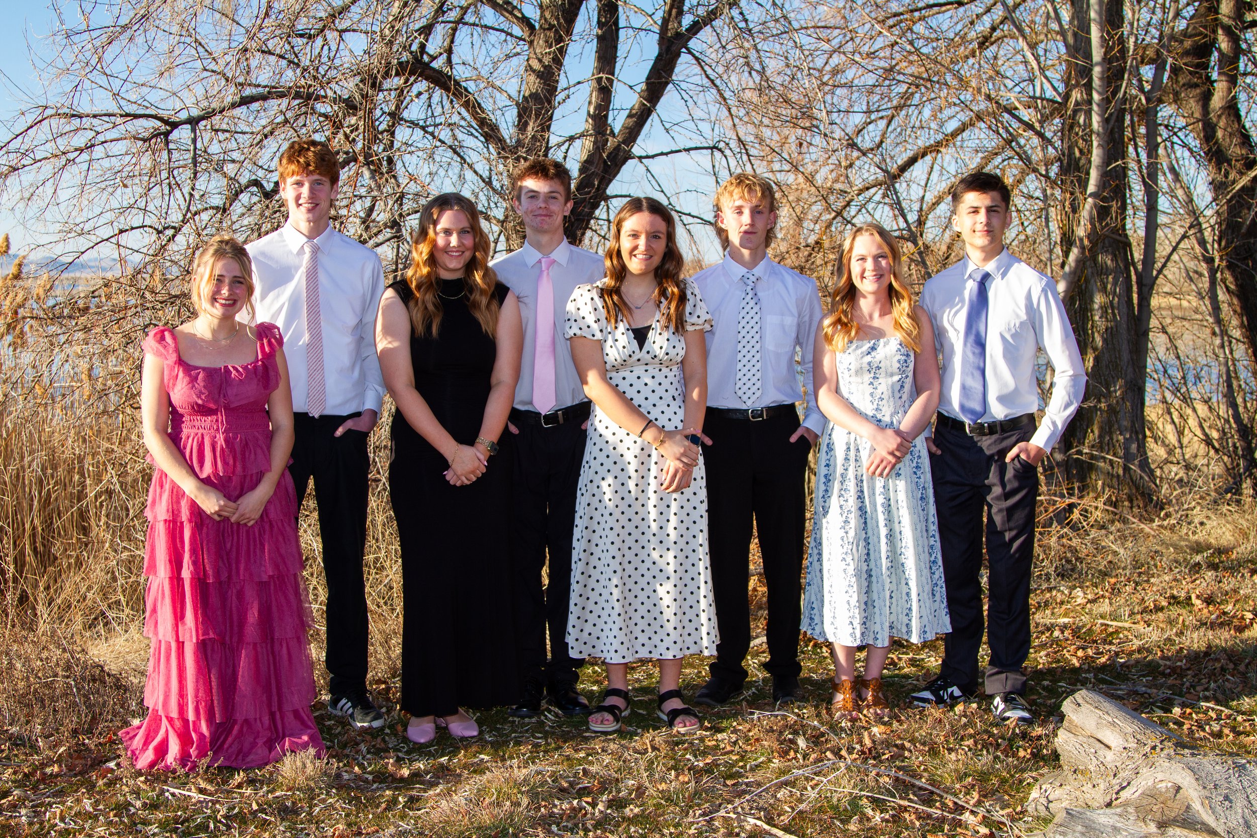 Group of nine young people standing outdoors in front of trees with bare branches, some in dresses and others in button-up shirts and slacks, on a sunny day.