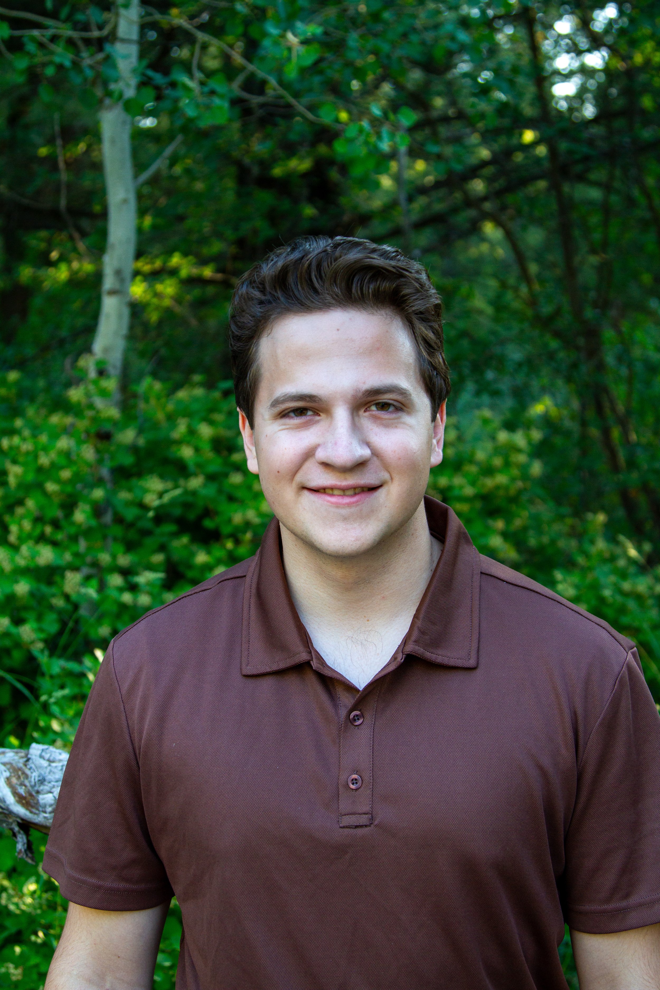 A young man smiling outdoors, wearing a brown polo shirt, with a background of green foliage and trees.