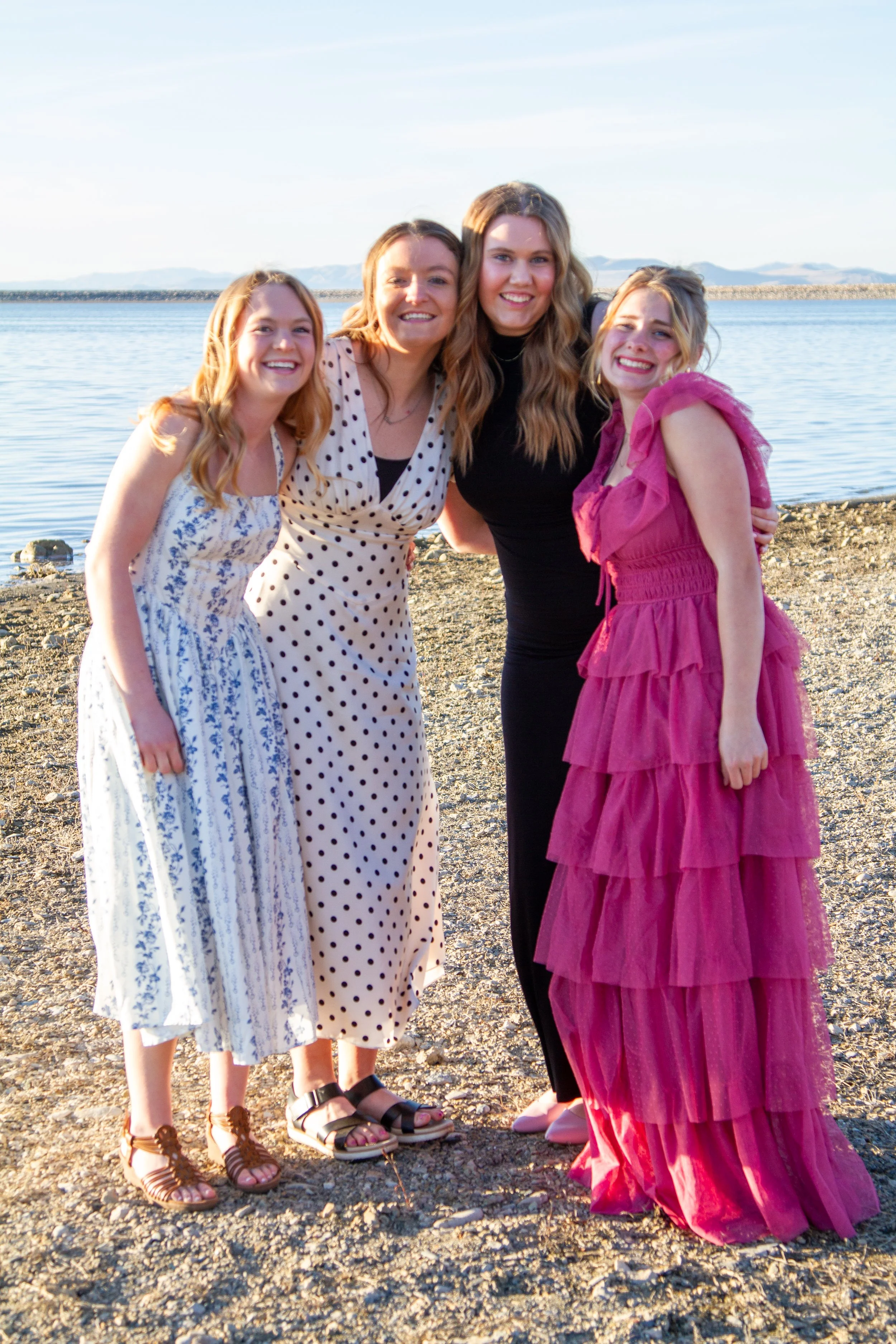 Four women standing on a rocky beach near a body of water, smiling and posing for the photo.