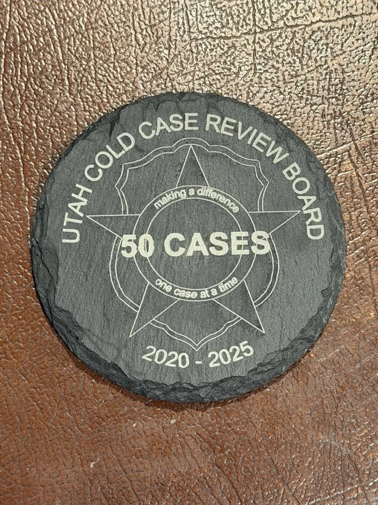 A round black coaster with white text reading "Utah Cold Case Review Board," "50 Cases," "2020 - 2025," and the phrase "making a difference one case at a time" inside a star graphic. The coaster is on a brown textured surface.