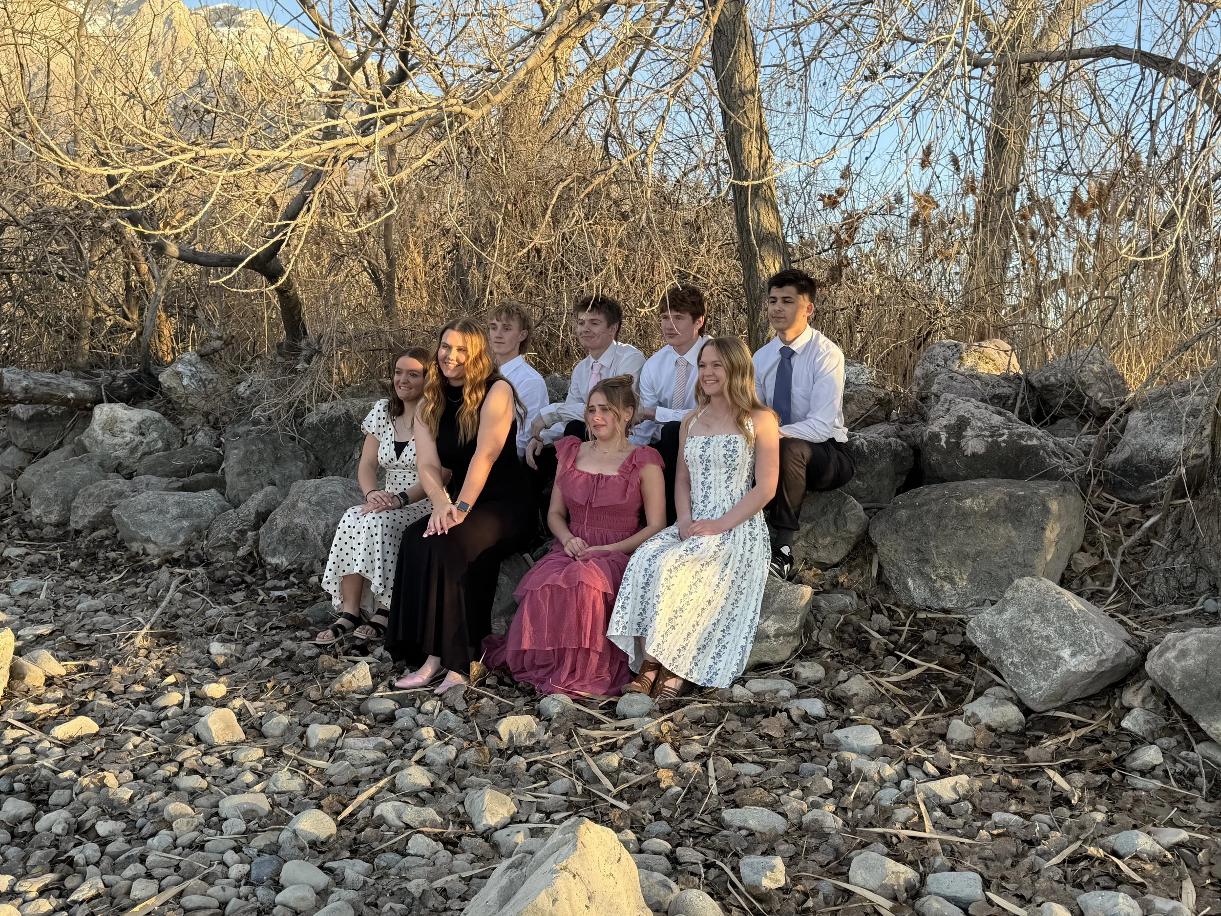 A group of eight teenagers dressed in formal attire sitting on large rocks in a wooded outdoor area during sunset.