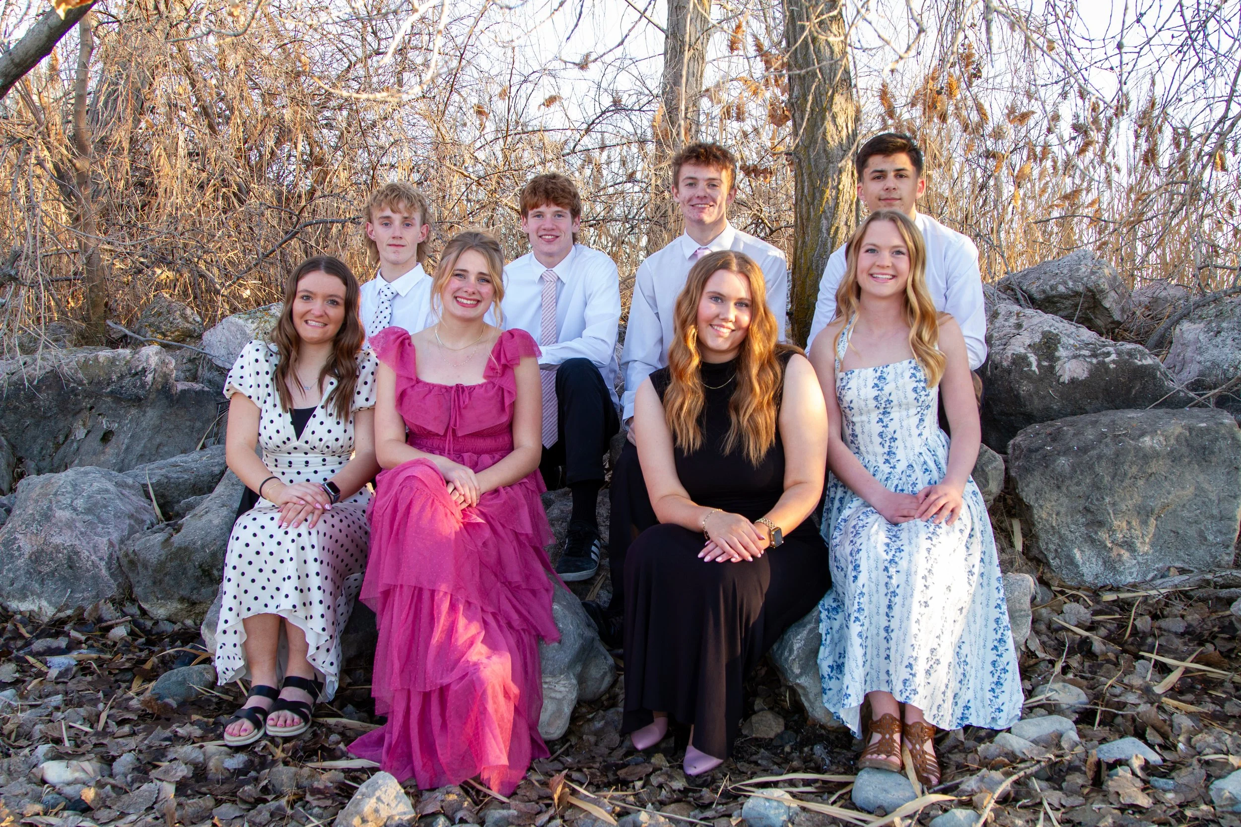 Group of nine teenagers, six girls and three boys, sitting on rocks outdoors during late fall or early winter, with bare trees and dry leaves around.