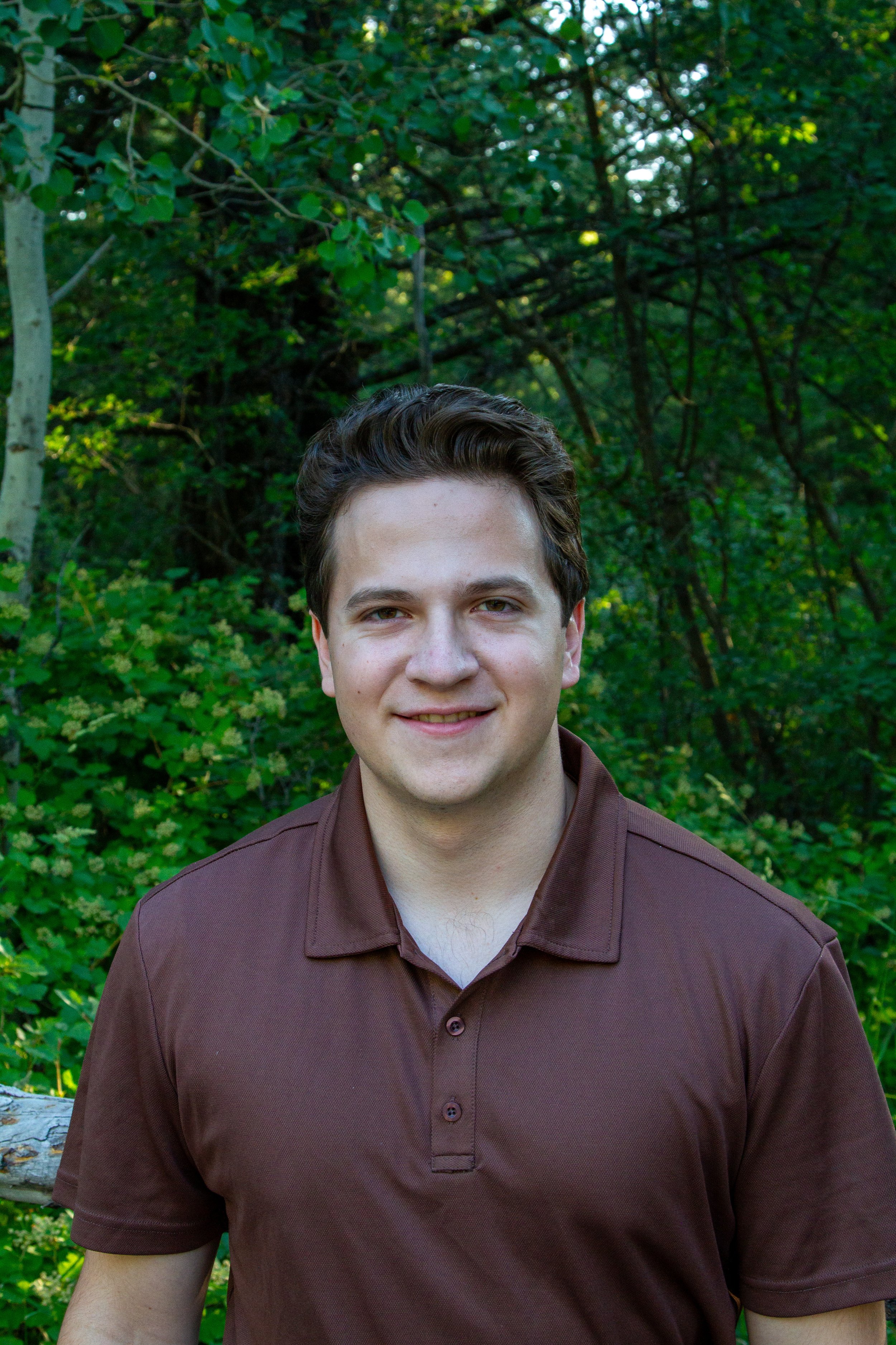 A young man with short brown hair smiling outdoors in front of green trees and foliage, wearing a brown collared shirt.
