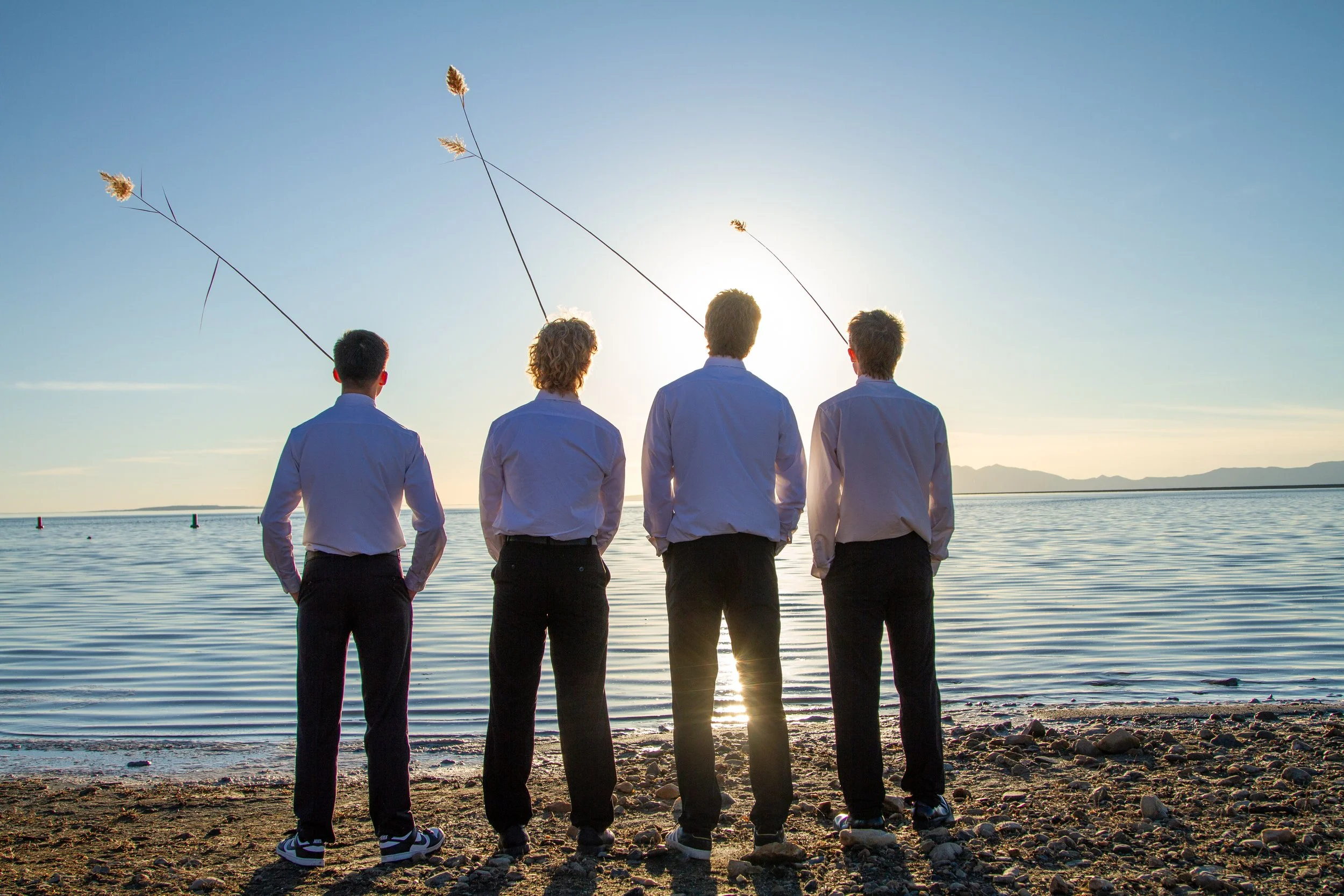 Four men in dress shirts and pants stand on a rocky beach facing water, fishing with rods as the sun sets in the background.