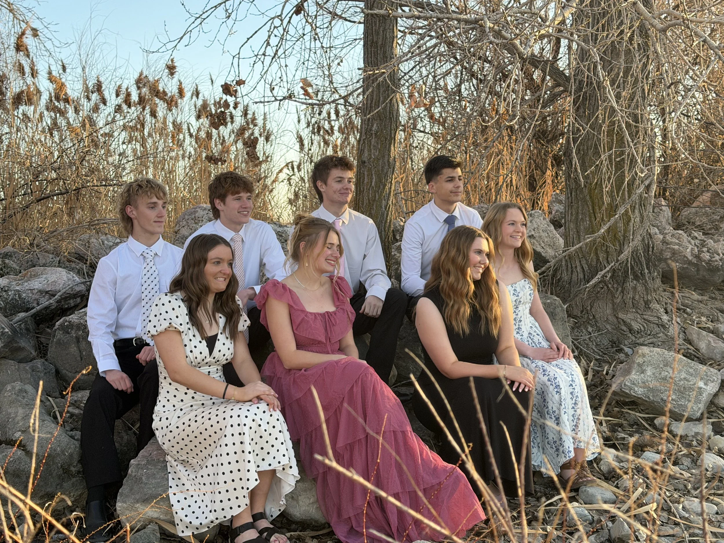 A group of eight young people, four males and four females, sitting on rocks outdoors during sunset, dressed in semi-formal attire, with trees and dry grass in the background.