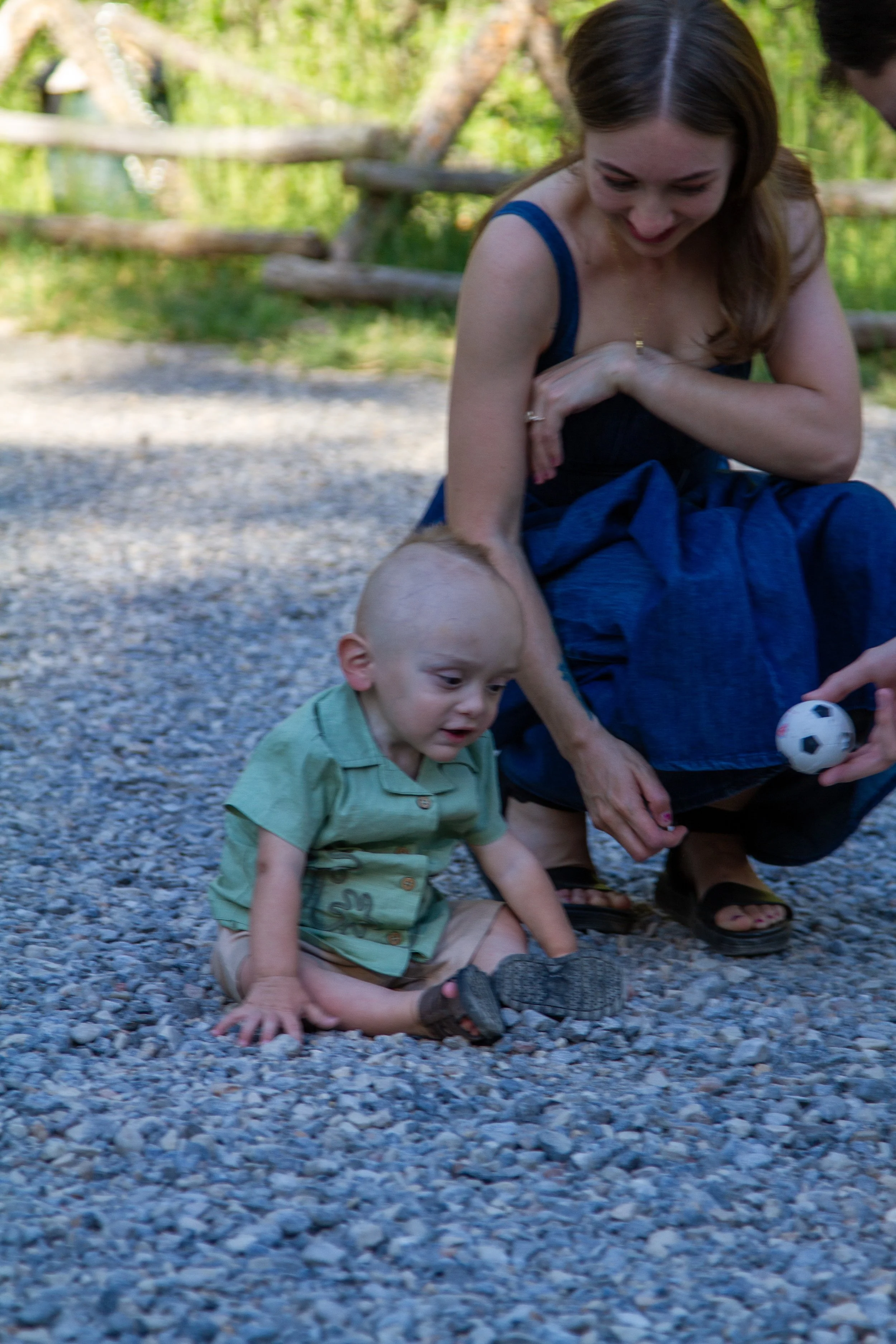 A woman and a young boy sitting on gravel outdoors, playing with a small soccer ball, with a wooden fence and green trees in the background.