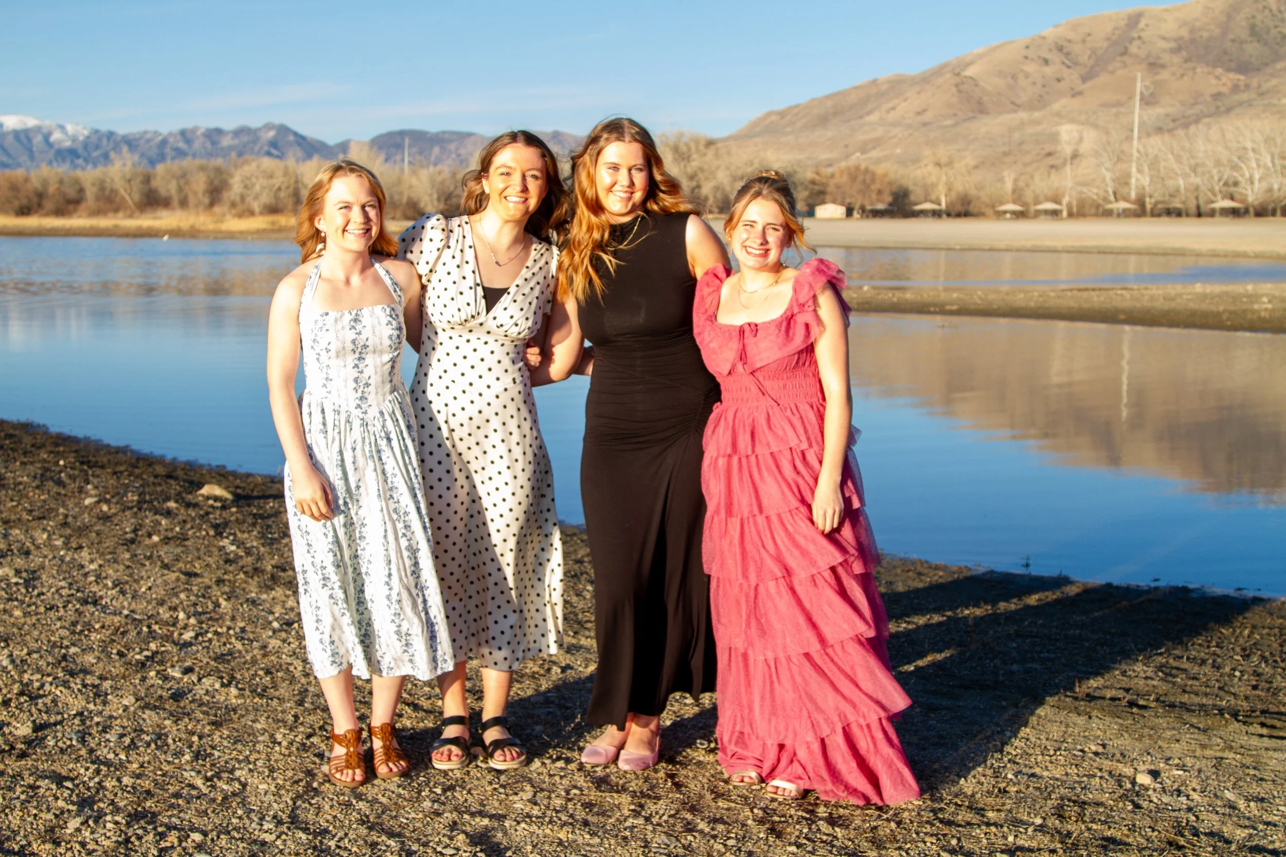 Four women in dresses standing by a lake with mountains in the background, smiling at the camera.