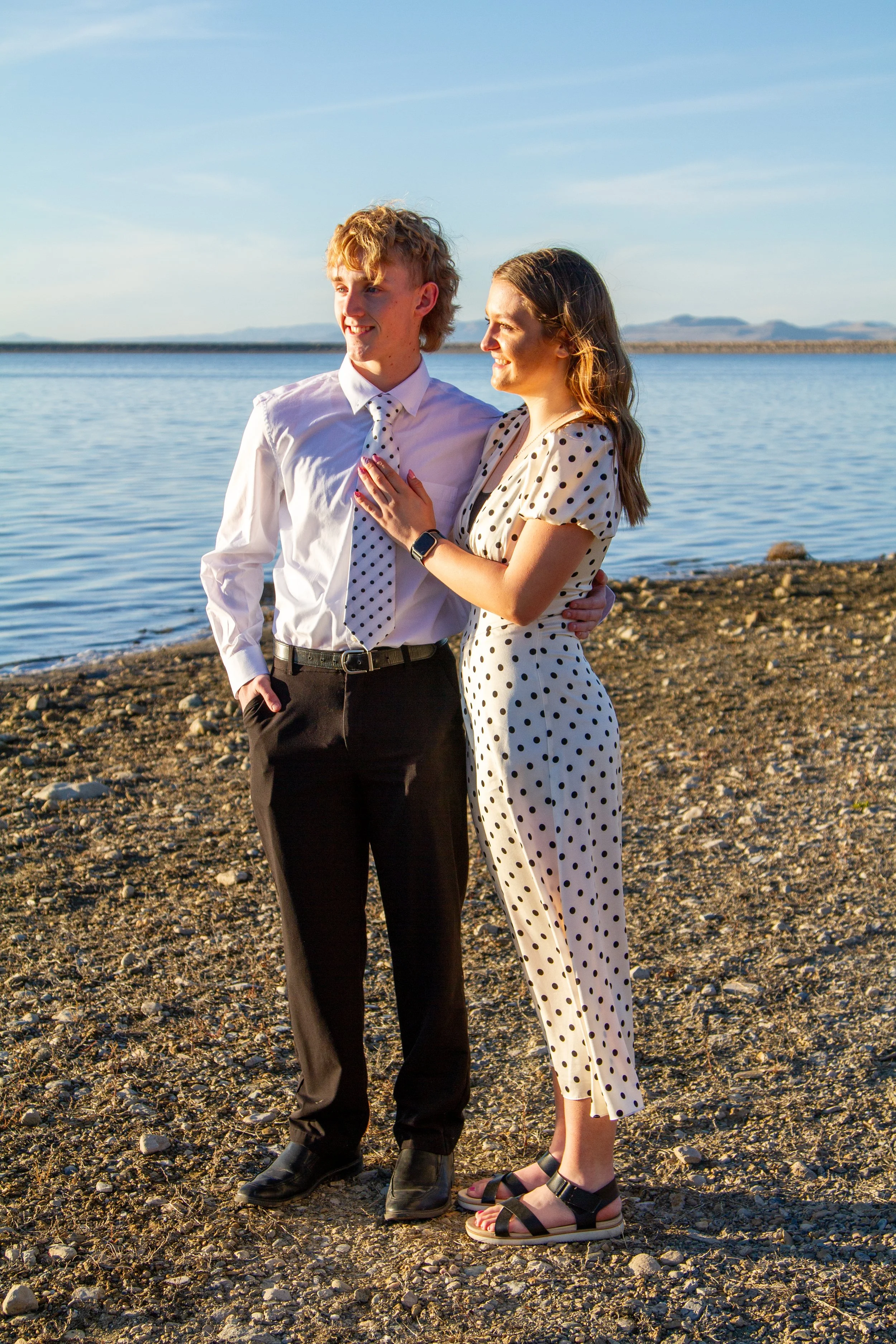 A young couple standing on a rocky beach near a body of water, with a sunset or sunrise sky in the background.