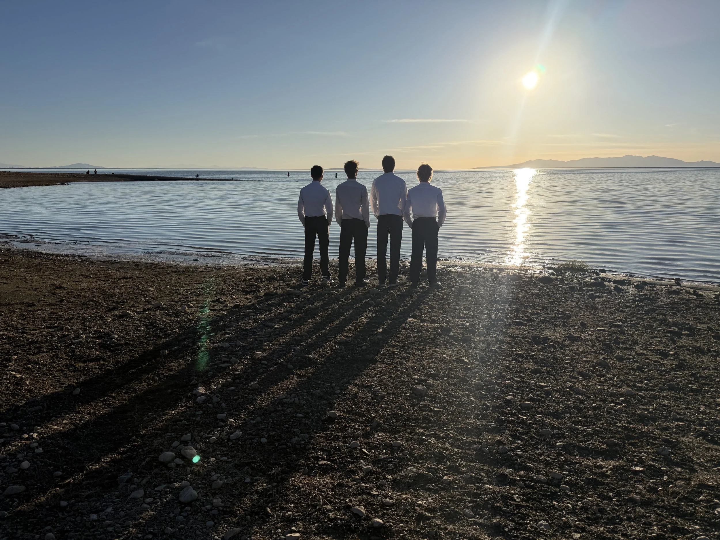 Four men in white shirts and dark pants stand on a rocky beach, facing the water during sunset, casting long shadows.