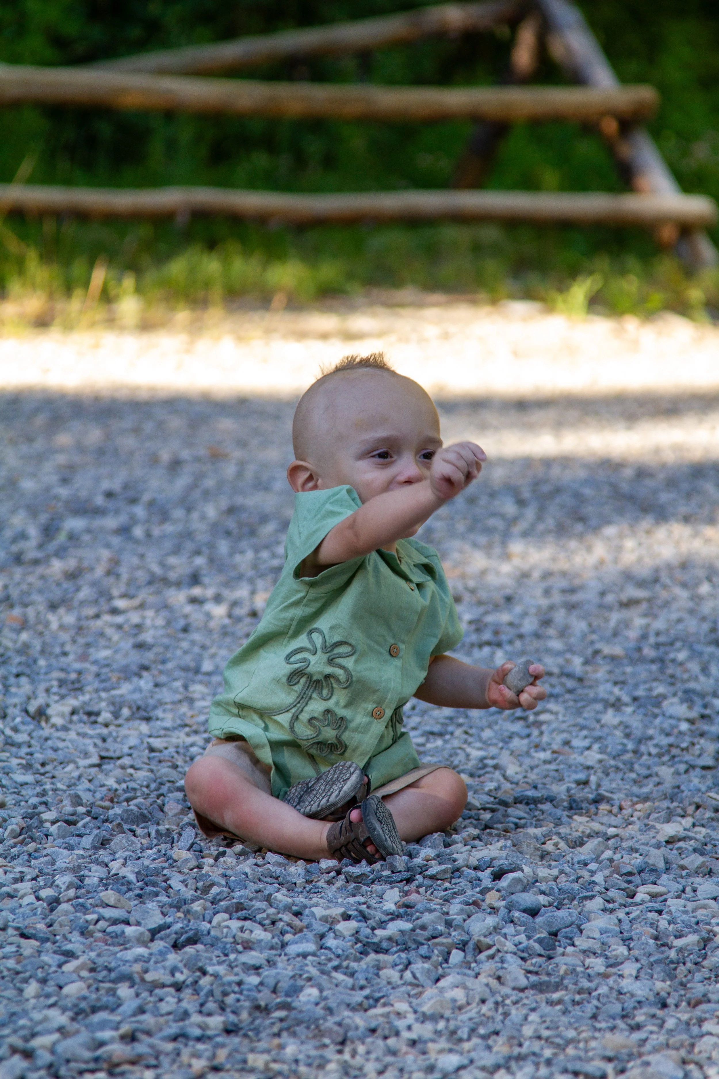 A young boy with a young child with a shaved head and light skin, sitting on a gravel surface outdoors. The boy is smiling and pointing, wearing a light green shirt with a flower design, brown shorts, and sandals. There is a wooden structure in the b
