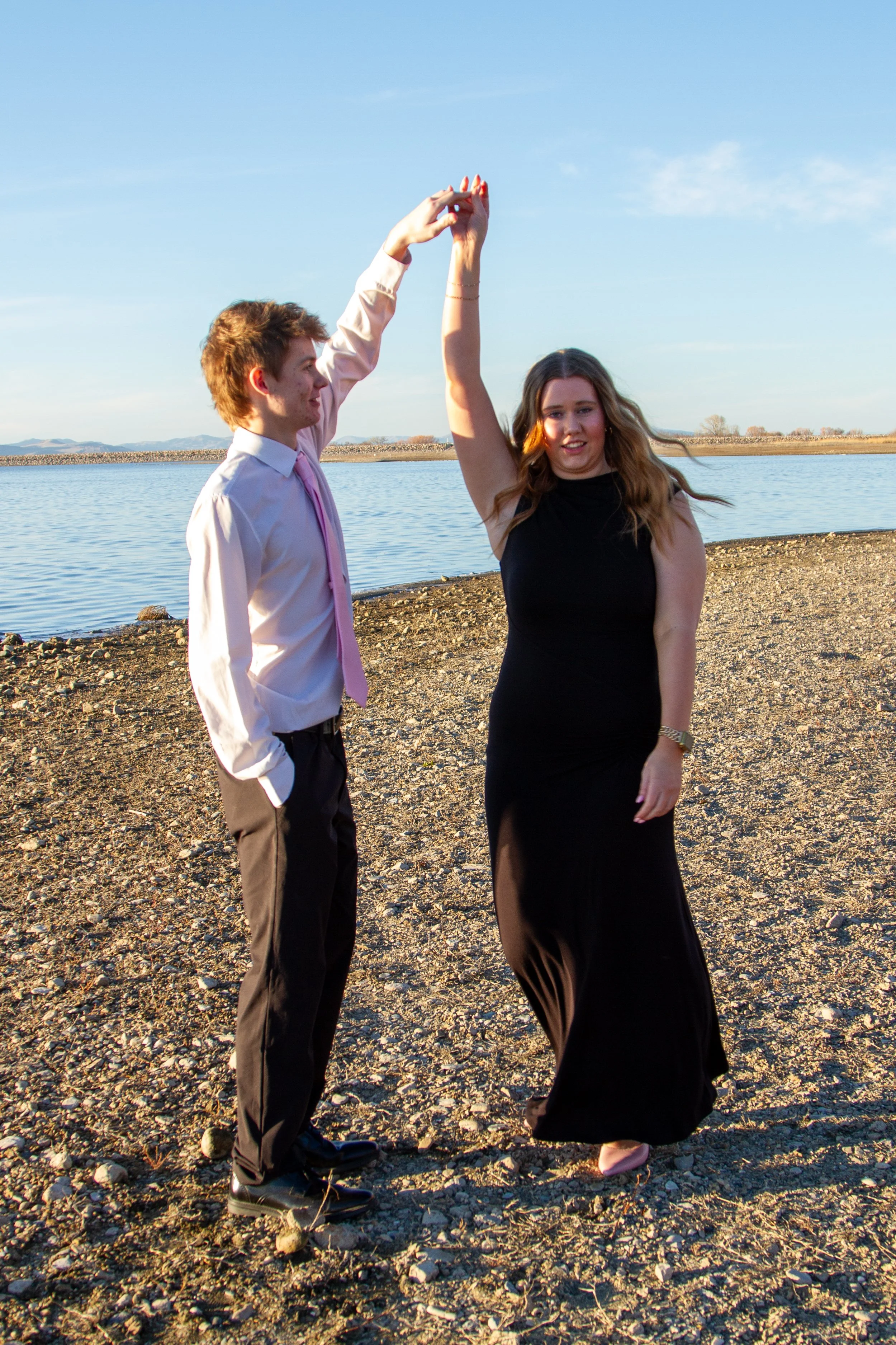 A young man and woman dance together outdoors near a body of water, holding hands overhead, with a clear sky in the background.