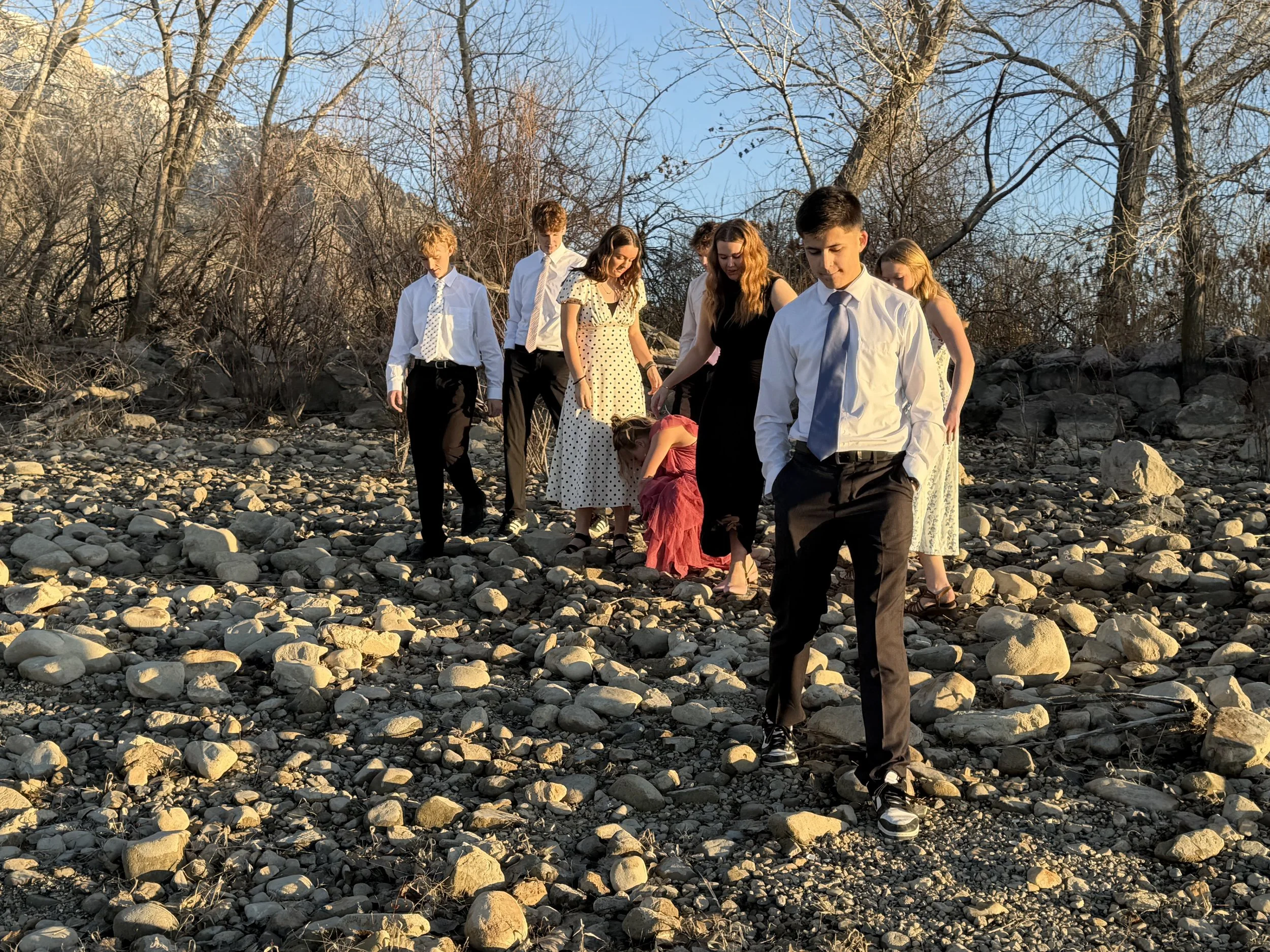 A group of people, mostly young, dressed up in semi-formal clothes walking on a rocky outdoor trail during sunset.