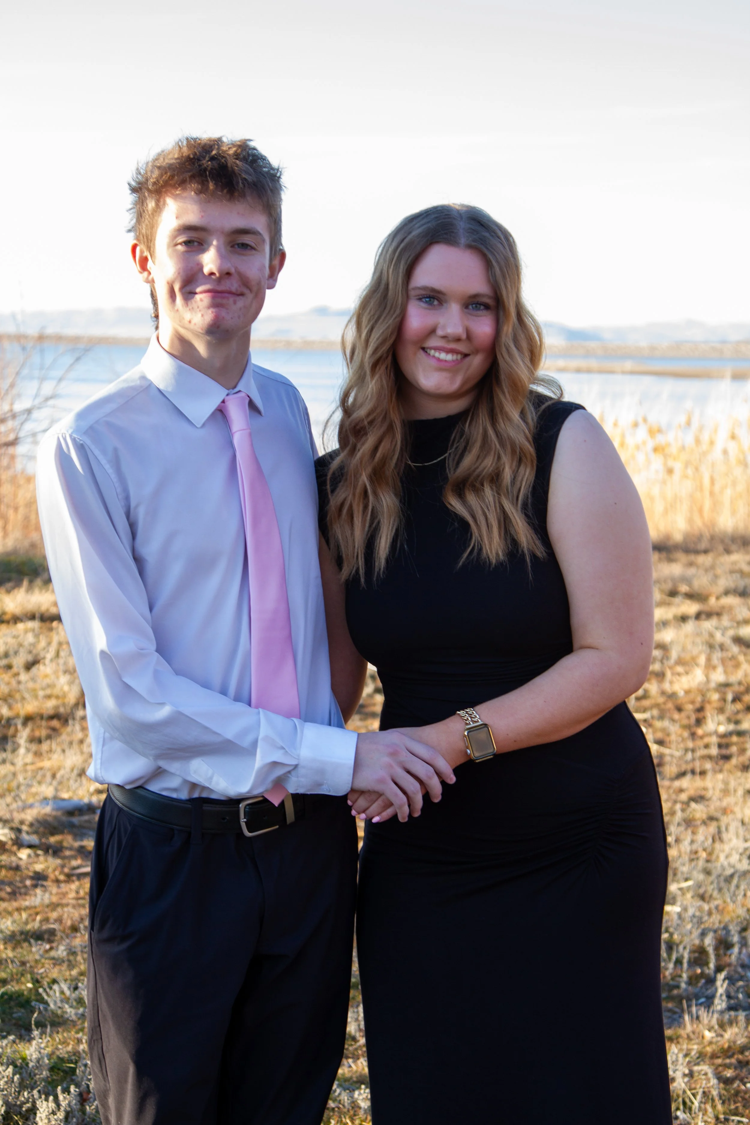 A young man and woman standing outdoors near a body of water with tall grasses in the background, holding hands and smiling at the camera.