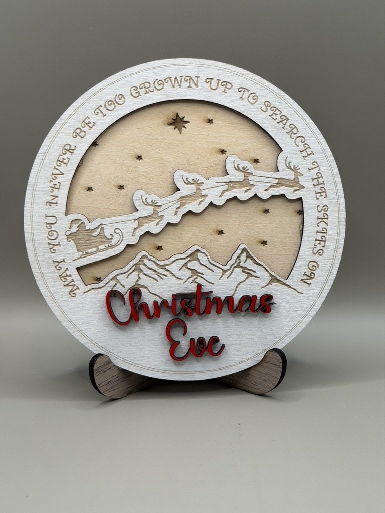 A decorative Christmas themed wooden piece with a mountain and Santa flying in his sleigh, surrounded by stars and a phrase about growing up to see stars. The words "Christmas Eve" are in red lettering at the bottom.