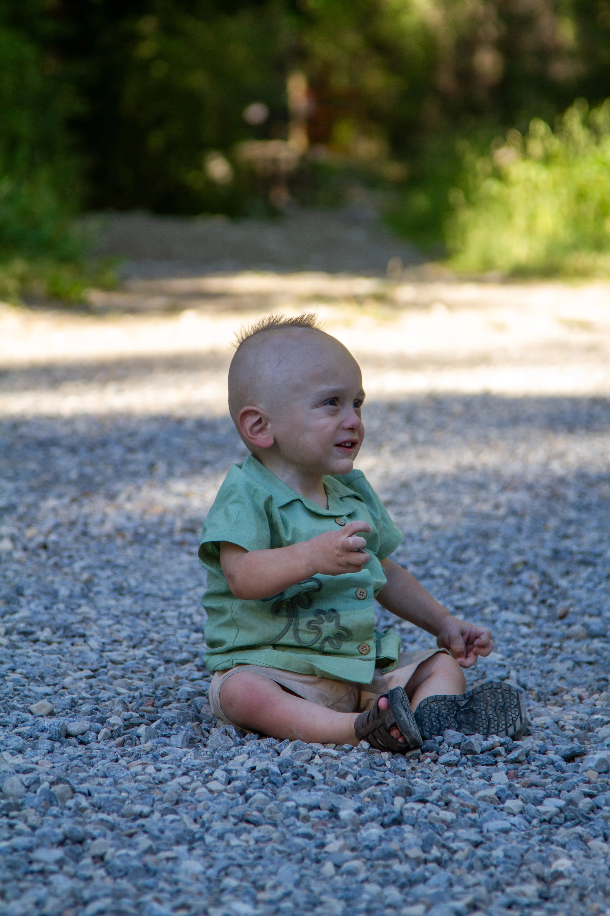A young boy with a shaved head sitting on a gravel surface outdoors, wearing a light green shirt and khaki shorts, smiling and looking off to the side, with greenery and blurred trees in the background.