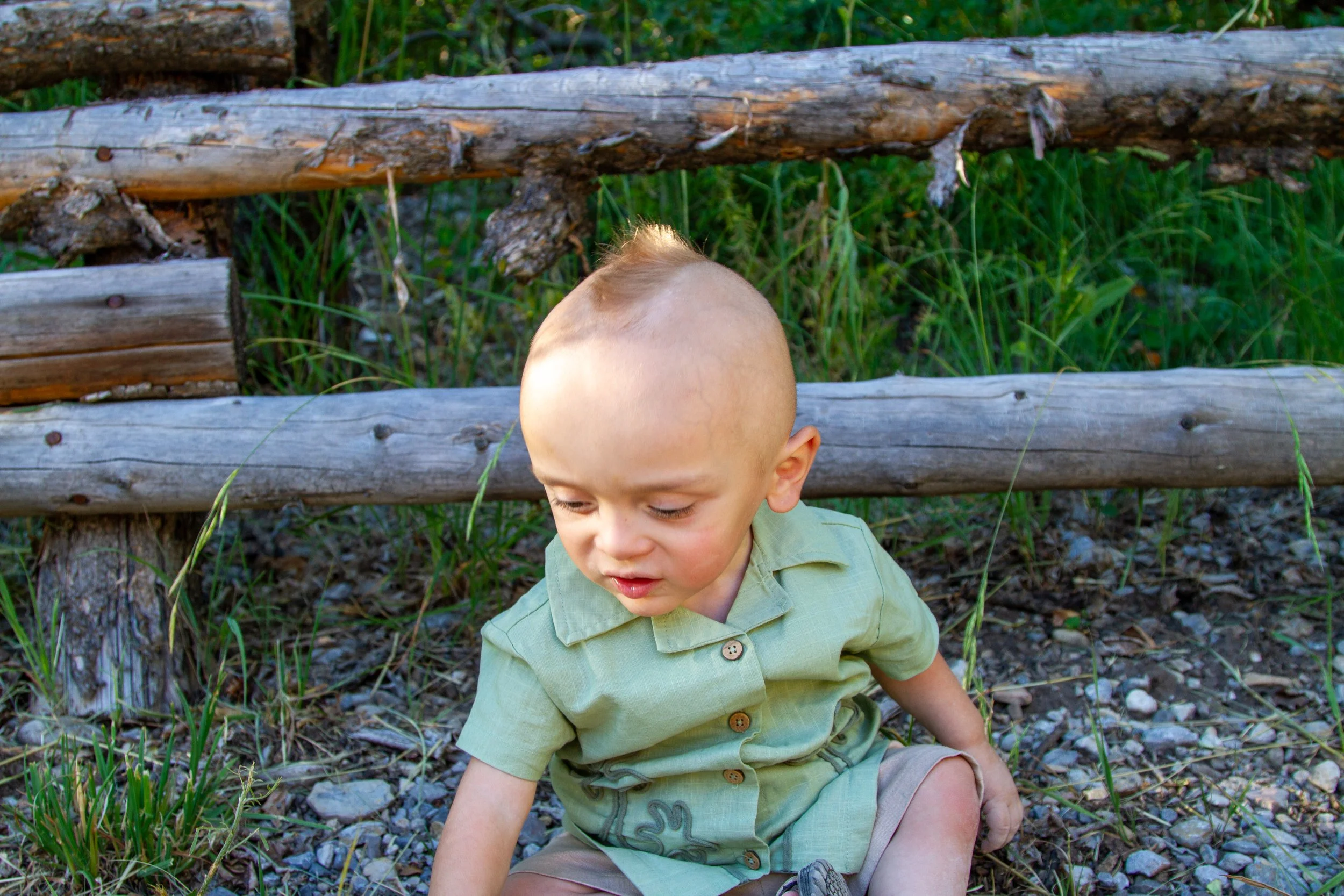 A young boy sitting outdoors near a wooden fence with green grass and rocks around him.