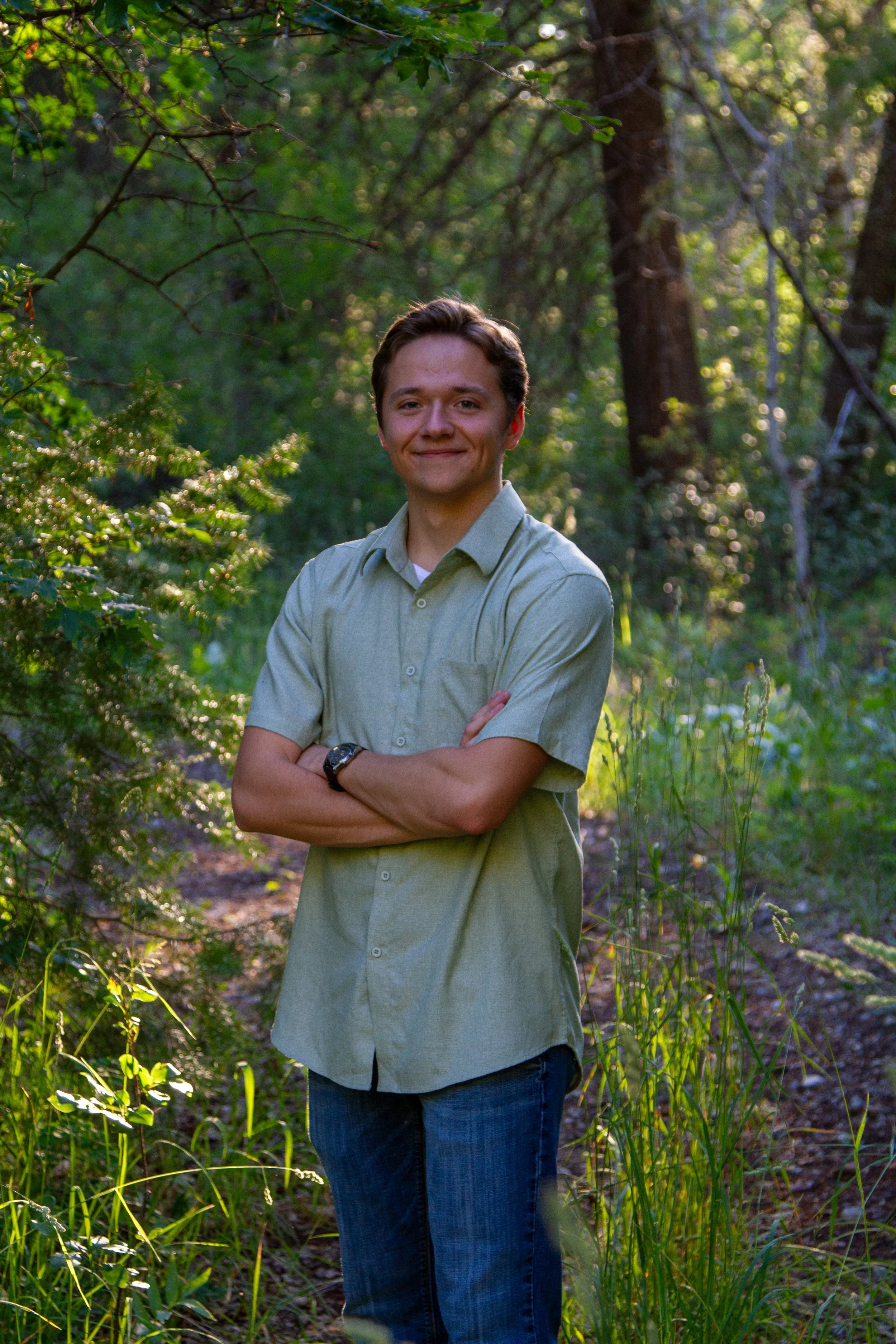 A young man standing outdoors in a wooded area with sunlight filtering through the trees, arms crossed, smiling