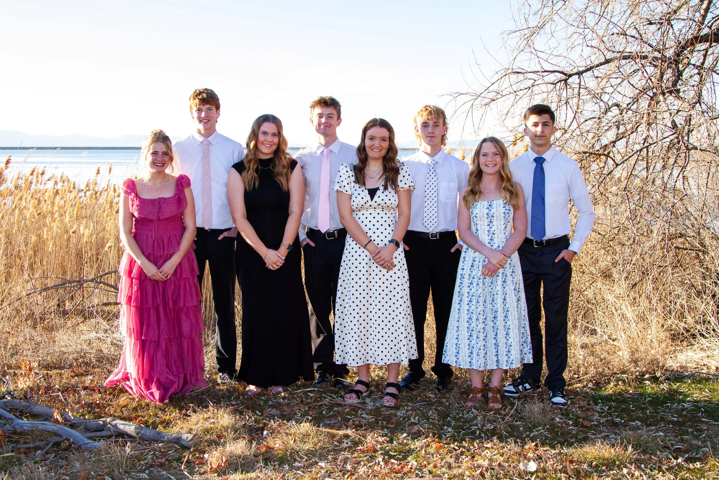 Group of nine young people standing outdoors in front of a body of water and a tree with no leaves, all dressed in semi-formal attire.