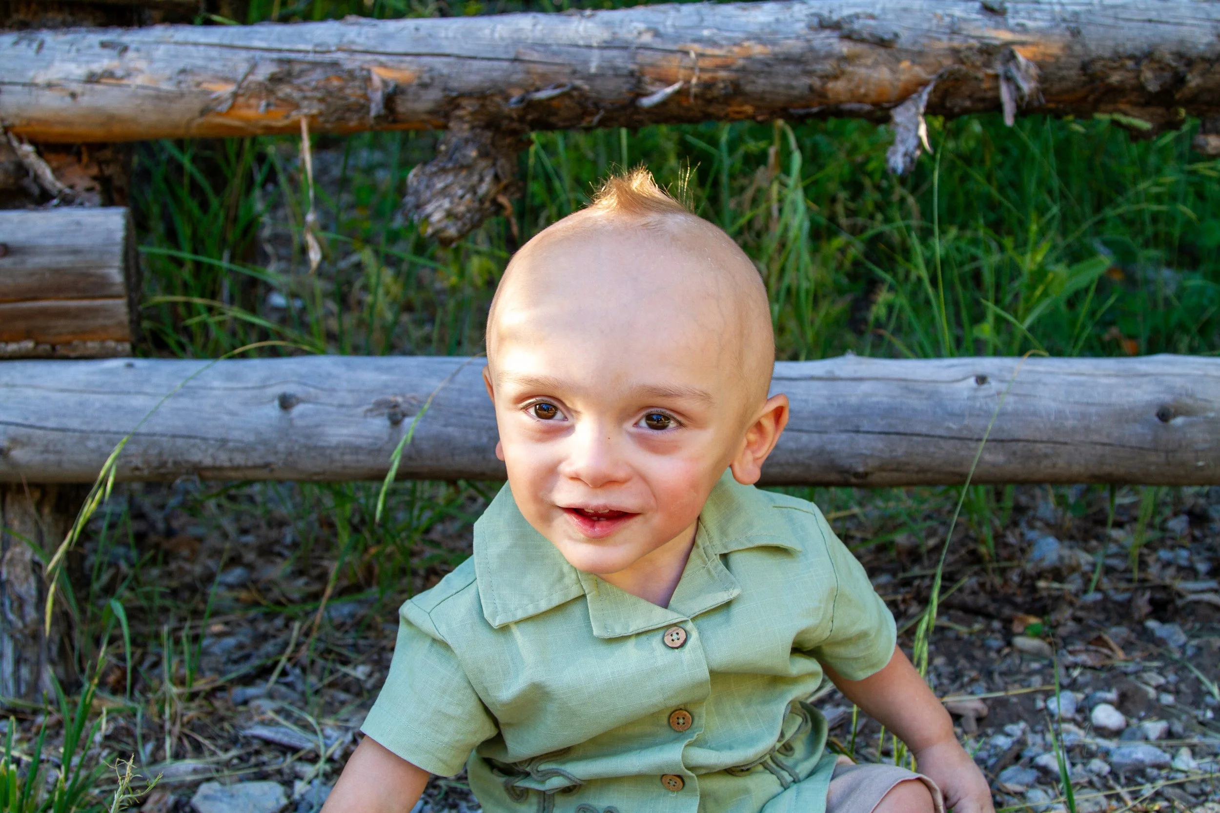 A young boy with a shaved head, wearing a light green shirt, smiling while sitting outdoors in front of a wooden fence with green grass and plants behind him.