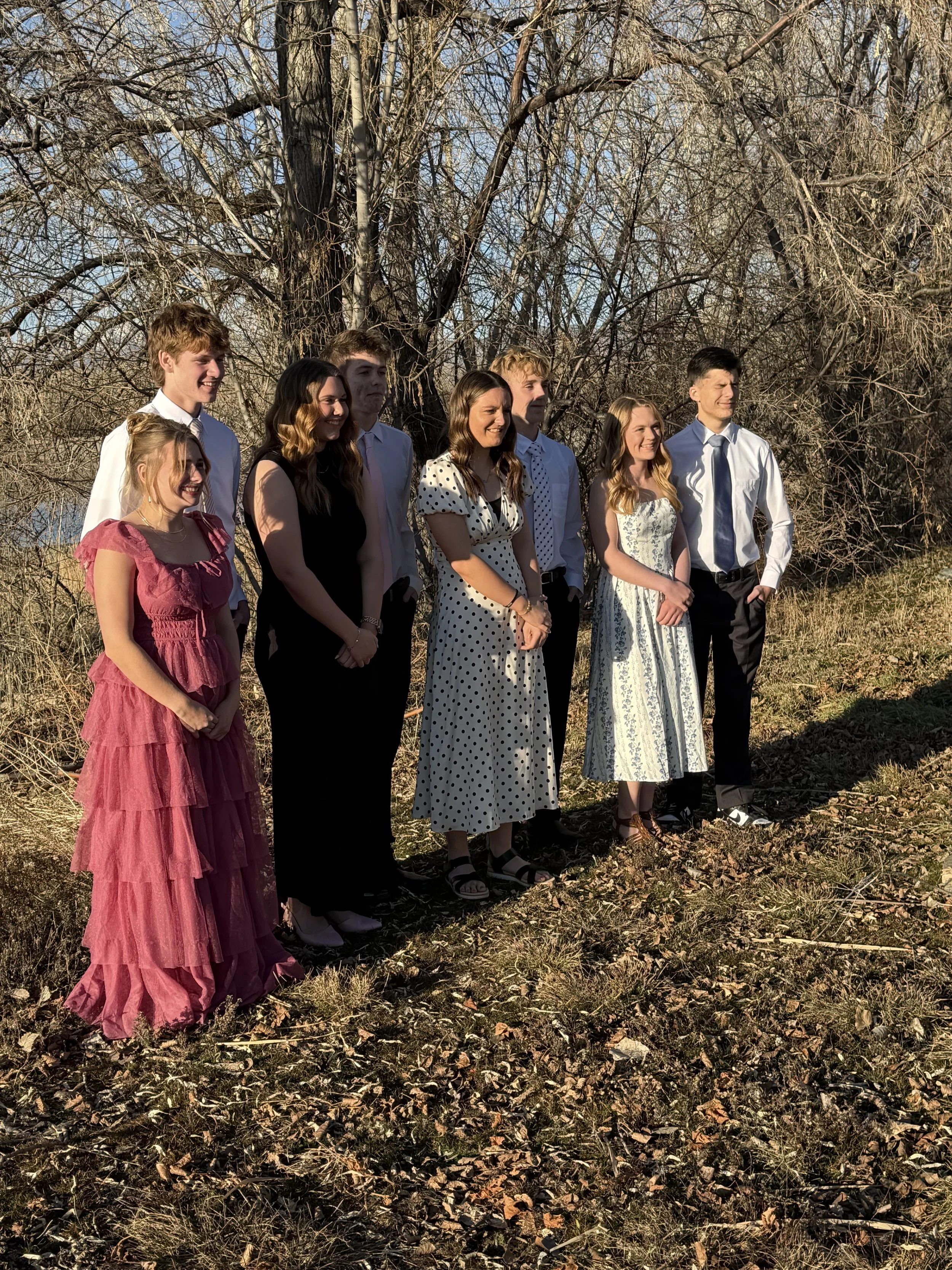 A group of seven young people standing outdoors on a grassy area with leafless trees in the background, dressed in semi-formal attire for a photo.