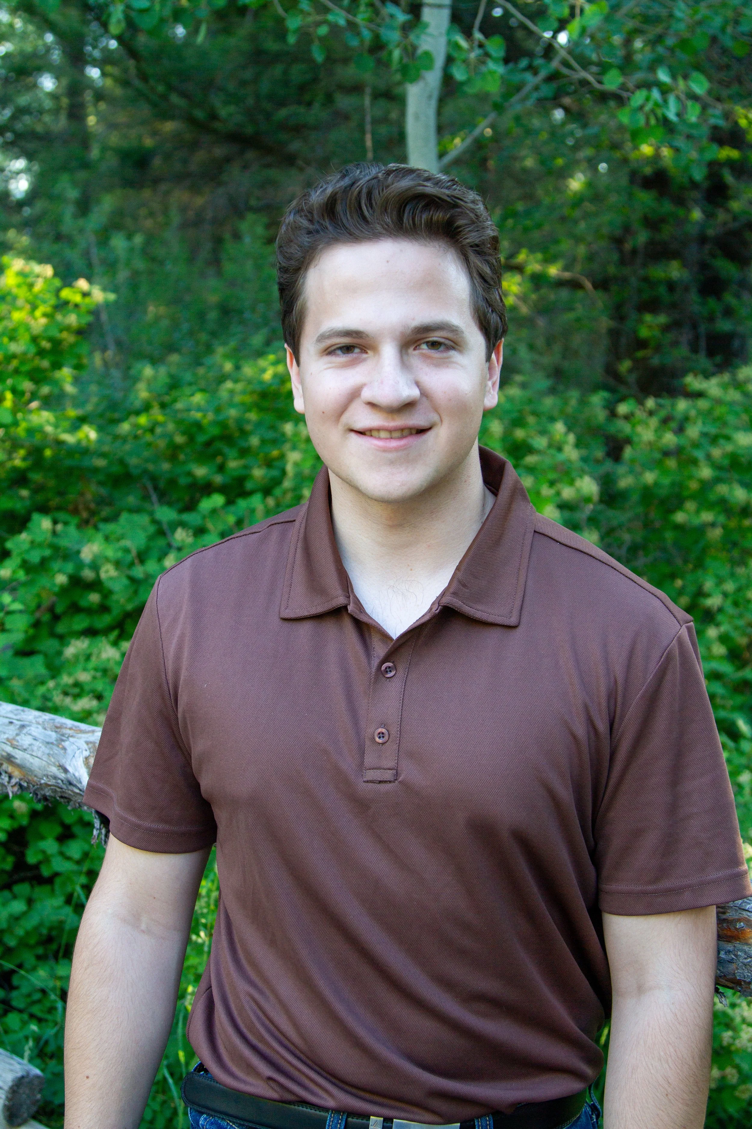 A young man with light skin, brown hair, and a slight smile, standing outdoors in front of green foliage, wearing a brown polo shirt and dark pants.