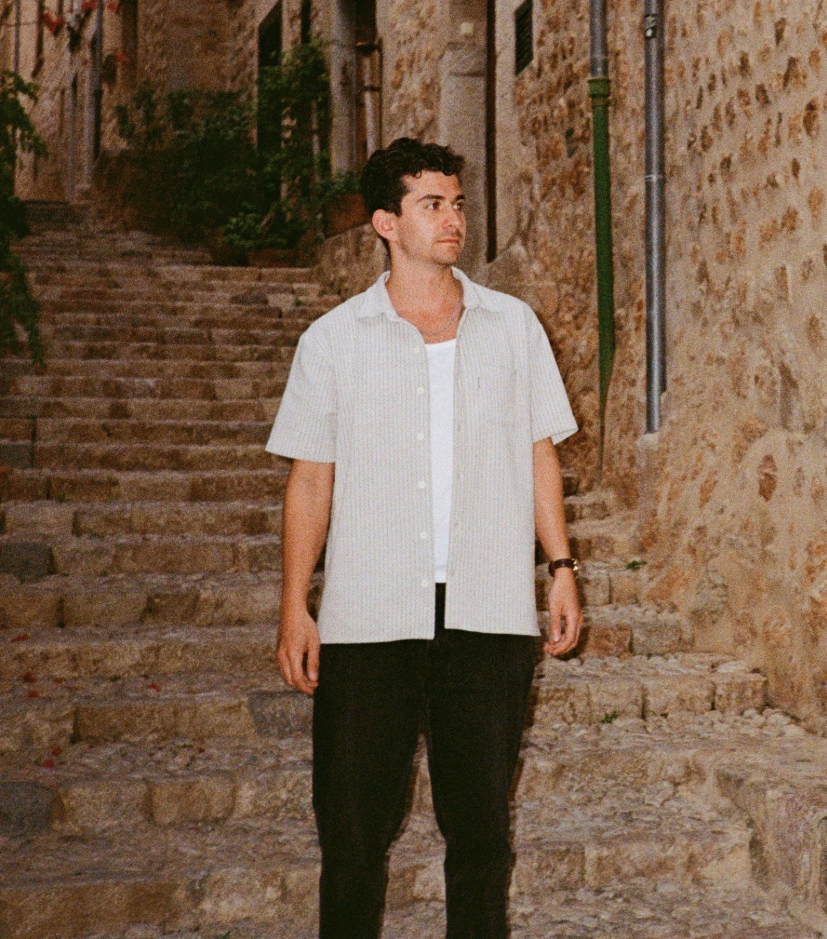 A man with dark hair, wearing a white short-sleeve button-up shirt over a white T-shirt, and black pants, standing on stone steps outdoors with a stone wall and windows in the background.