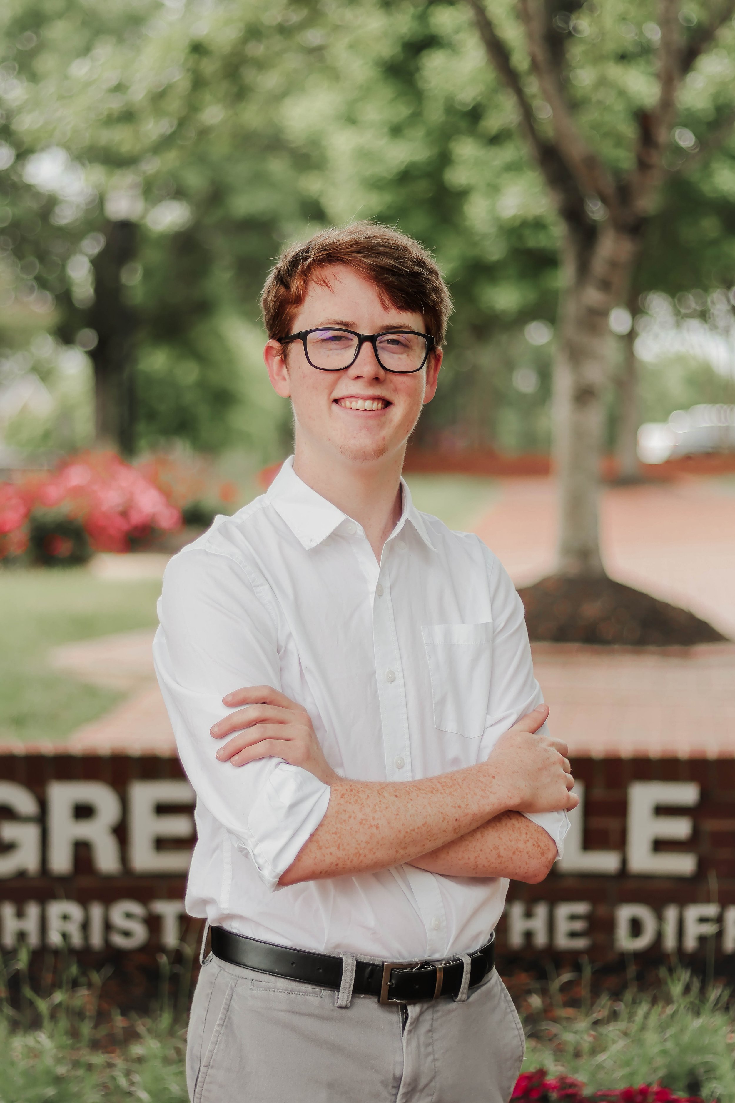 A young man with glasses and freckles wearing a white button-up shirt and beige pants, standing outside with folded arms, smiling at the camera.