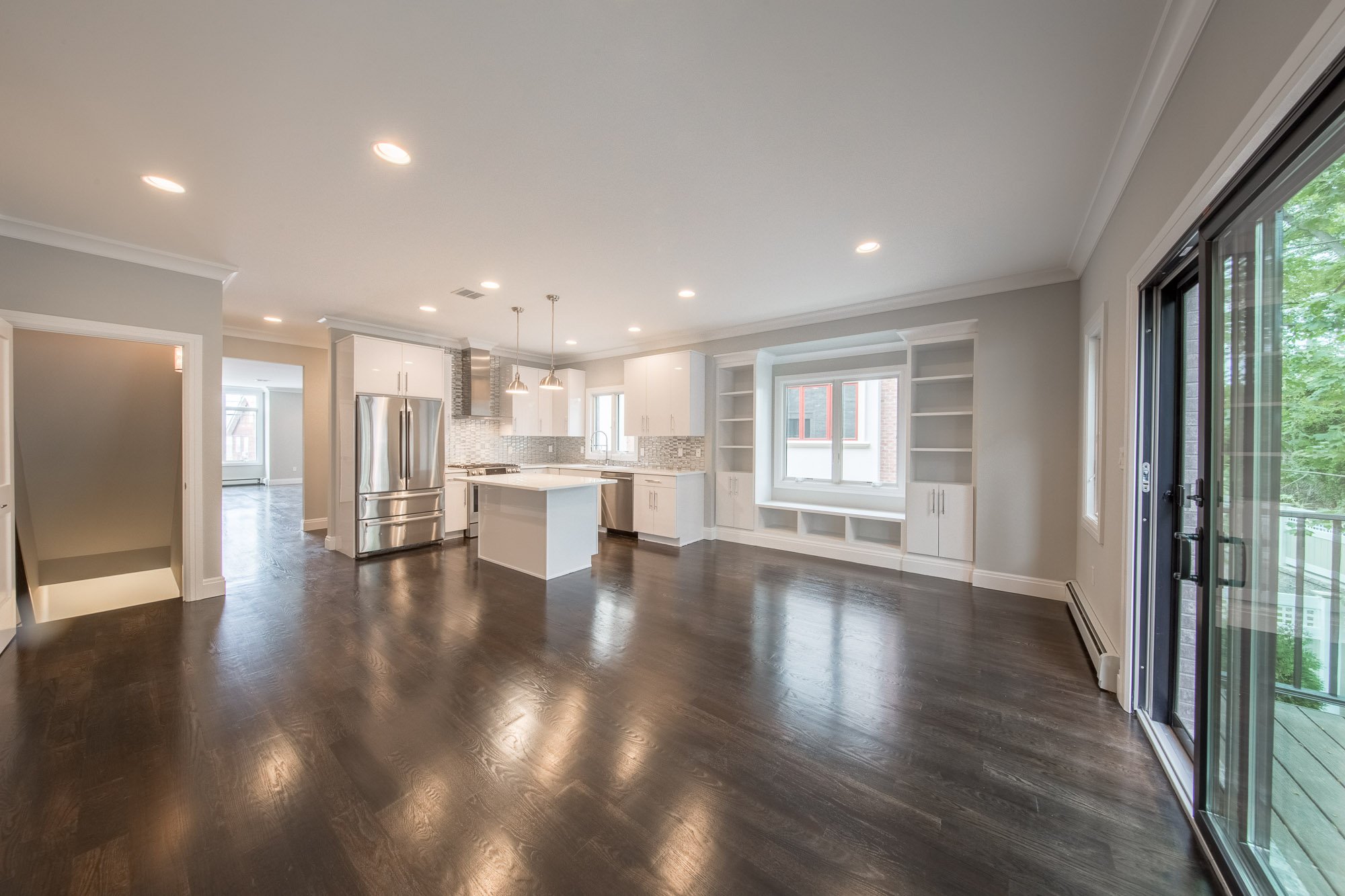 Modern kitchen with white cabinets, stainless steel refrigerator, island, and dark wood flooring, adjacent to a sliding glass door leading outside.