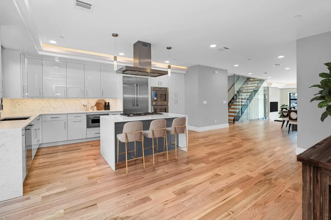 Open-concept modern kitchen with light gray cabinets, a white island with a dark gray base, three beige bar stools, and stainless steel appliances, including a double oven and a refrigerator, with a living area and staircase visible in the background.