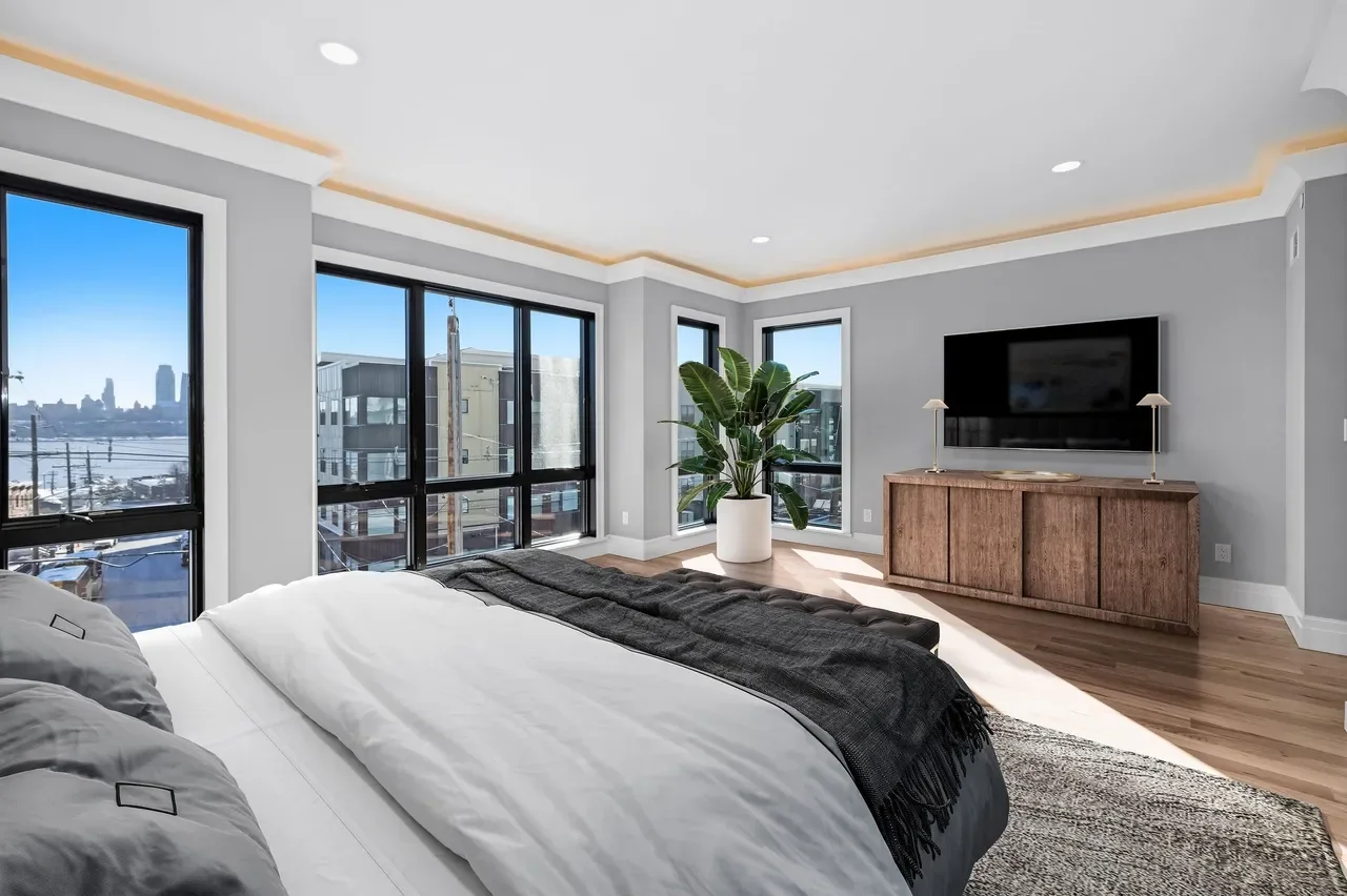 Modern bedroom with large windows overlooking a city skyline, a bed with white and gray bedding, a wooden dresser with a mounted flat-screen TV, a large green plant in a white pot, and hardwood floors.