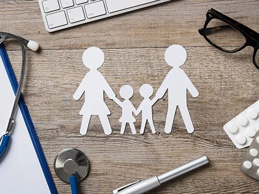 Paper cutouts of a family holding hands on a wooden desk surrounded by medical and office supplies.