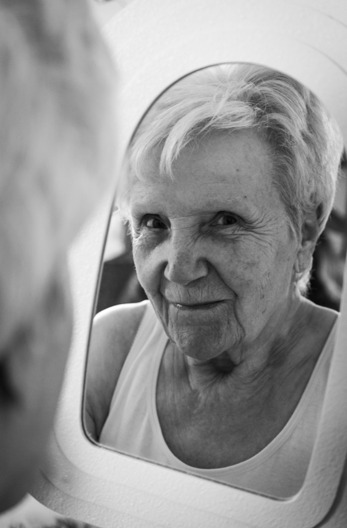 An elderly woman looking at her reflection in a mirror with a playful expression.