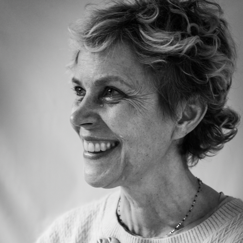 A smiling older woman with short curly hair, wearing a sweater and a necklace, touching her chest with one hand and wearing a ring.