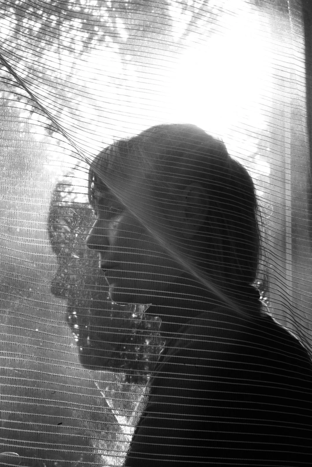 Silhouette of a woman with long hair looking out a window with a sheer curtain, sunlight streaming through, and trees visible outside.