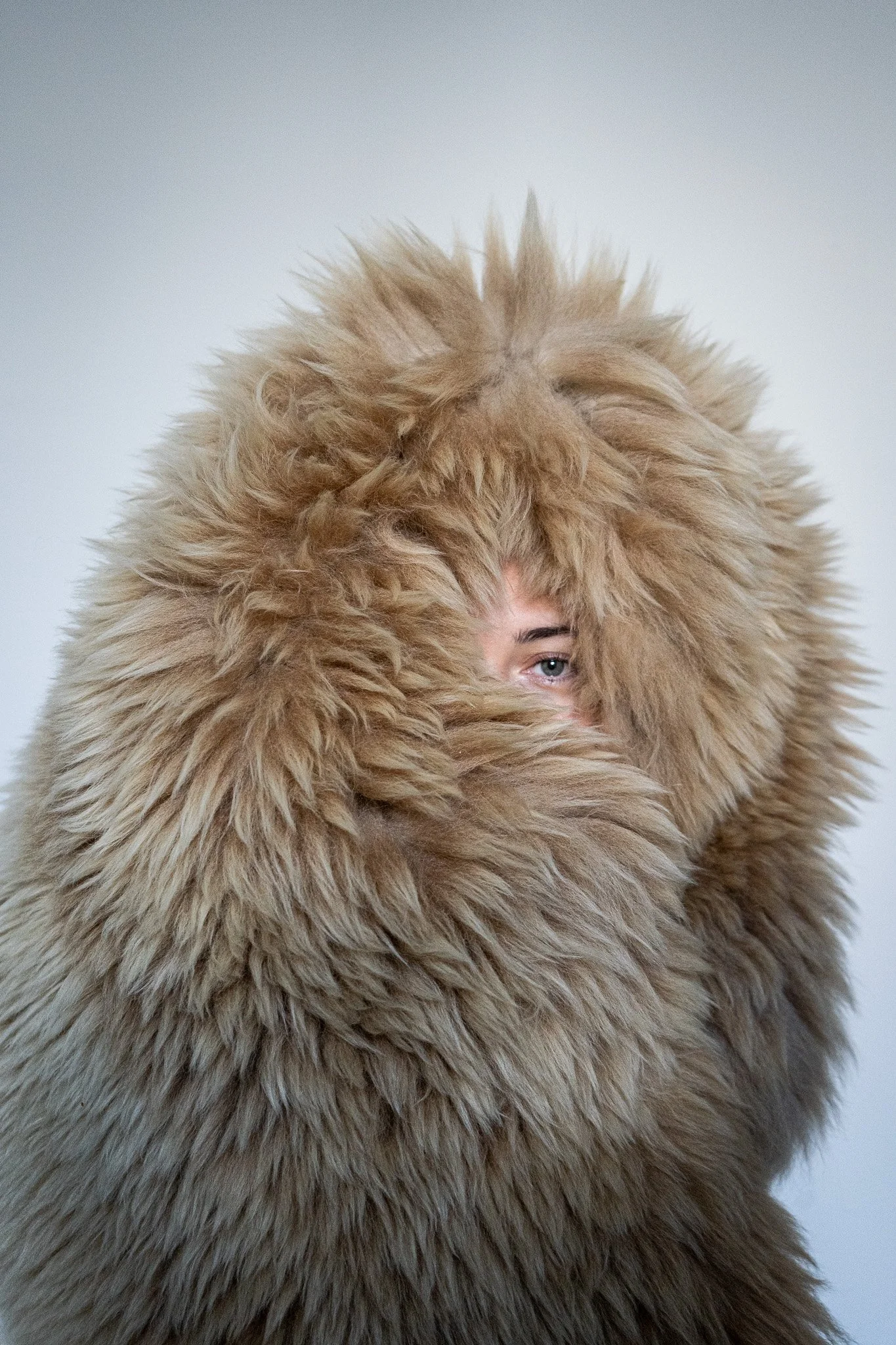 Person wrapped in a large, fluffy, tan fur garment, with only one visible eye, against a plain background.