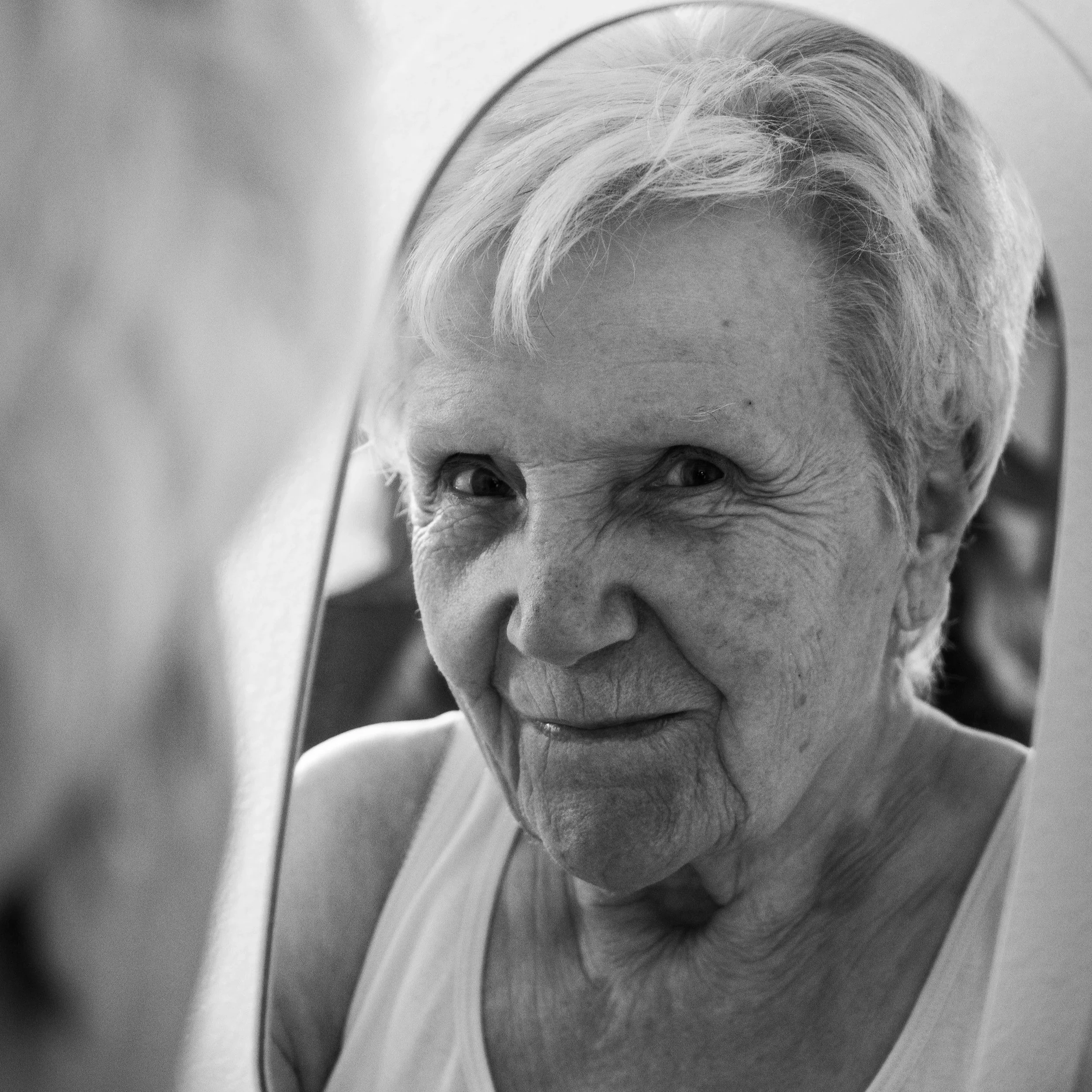 A smiling older woman with short curly hair, wearing a sweater and a necklace, touching her chest with one hand and wearing a ring.