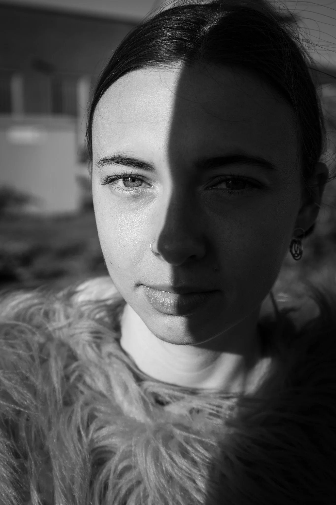 Black and white portrait of a woman with a partially shadowed face, wearing a fur coat and earrings, outdoors with blurred background.