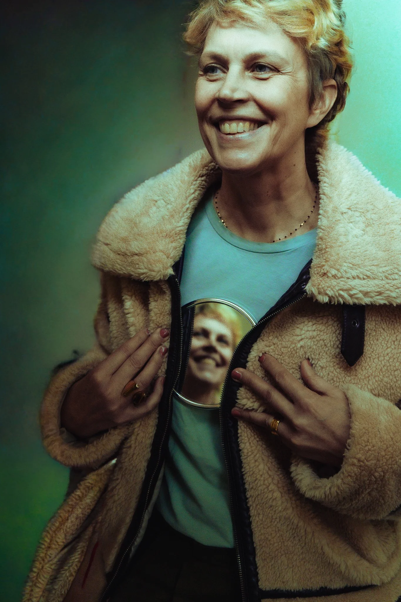 A woman with short blonde hair is smiling, wearing a beige fuzzy jacket over a light blue shirt. She is holding her jacket open to reveal a photograph of another smiling woman on her shirt.