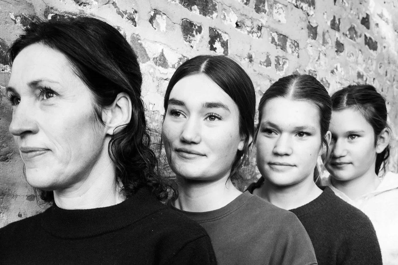 Four women standing in a line against a brick wall, facing to the left, in black and white.