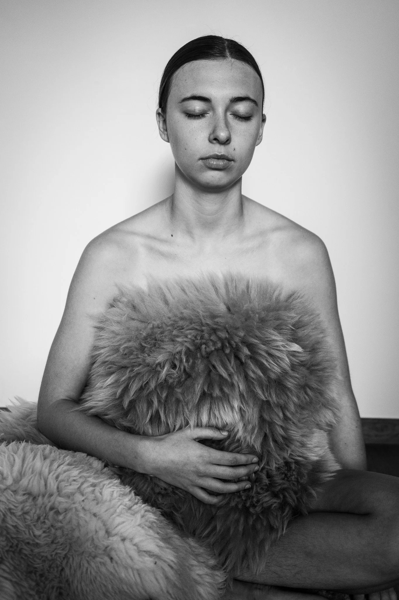 A black and white photo of a woman with her eyes closed, holding a large piece of furry fabric against her chest, with another piece of fur draped over her lap.