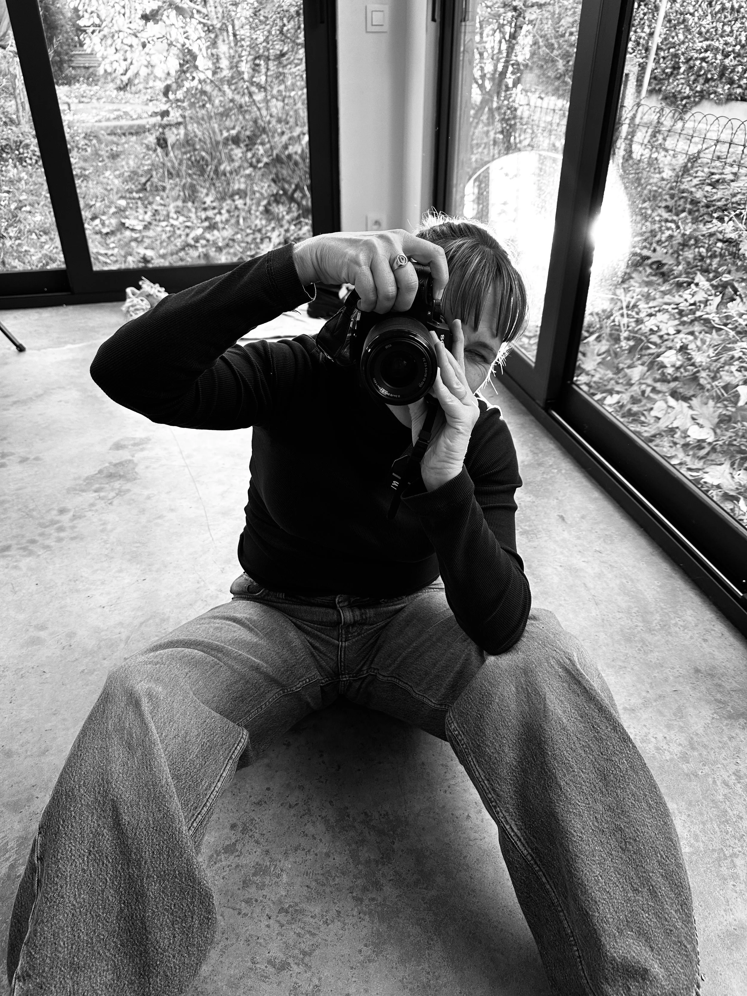 Person sitting on the floor in front of large glass windows, taking a photo with a camera, with outdoor foliage visible through the windows.