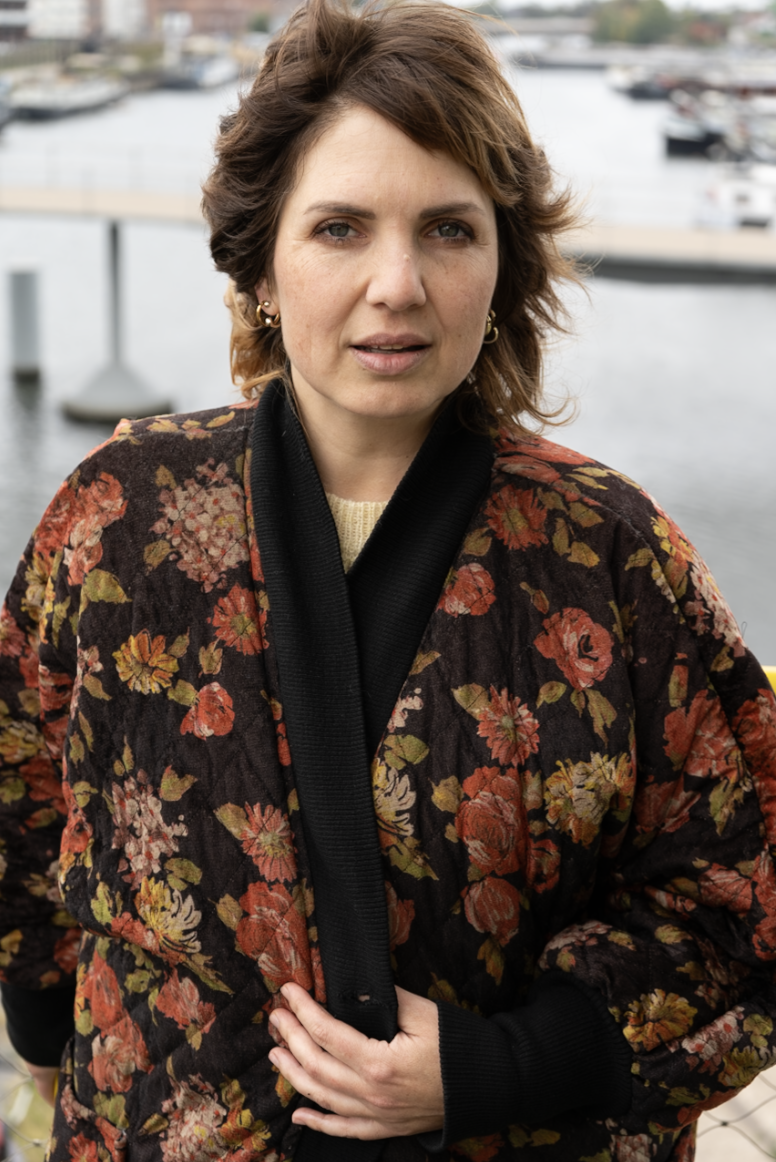 A woman with short brown hair wearing a black and floral patterned jacket standing near a river with boats in the background.