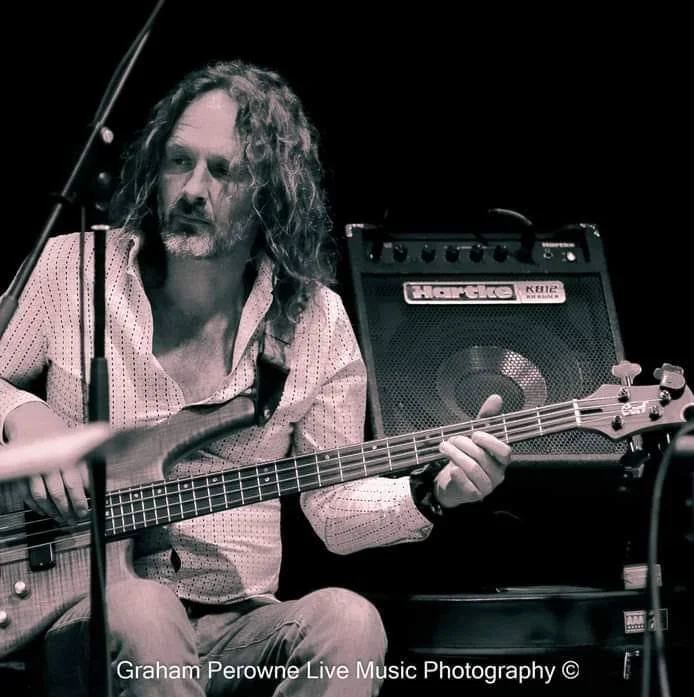 Man with long curly hair and beard playing a bass guitar in a live music setting, with a small amplifier in the background.