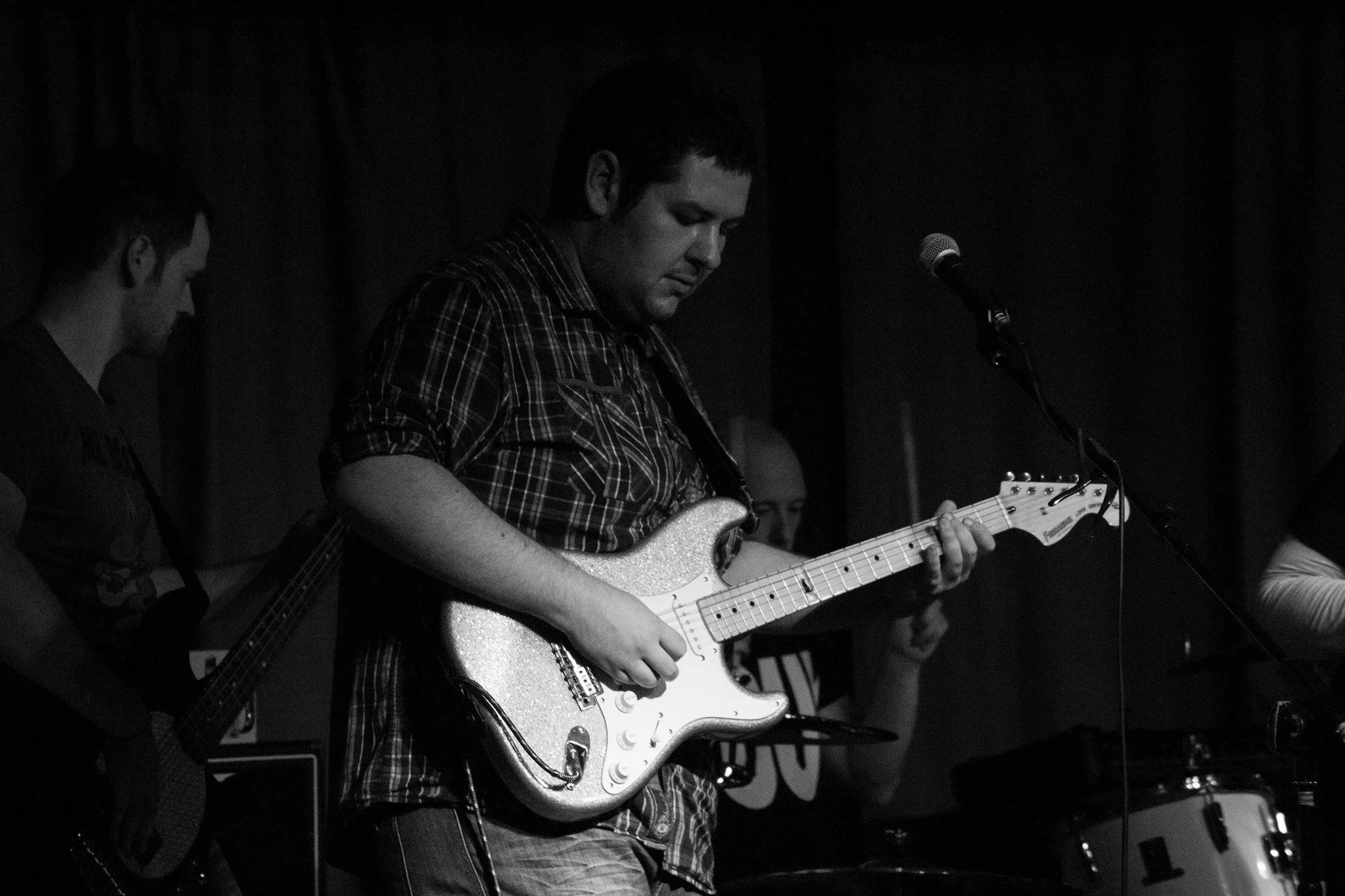 A group of musicians performing on stage. The main focus is on a man playing an electric guitar, wearing a plaid shirt and concentrating on his instrument. Other band members are visible in the background.