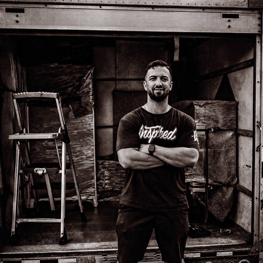 A man with a beard in a black T-shirt standing with arms crossed in an industrial setting, possibly inside a truck or storage container.