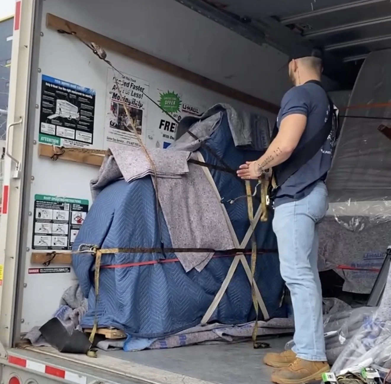 Man standing inside a moving truck, securing a large item wrapped in blankets and straps.